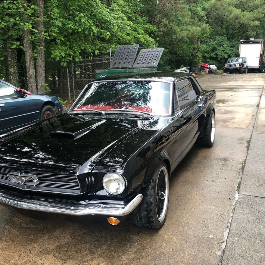 Black classic Mustang car, with chrome details, parked on a paved lot, with other vehicles and trees in the background.