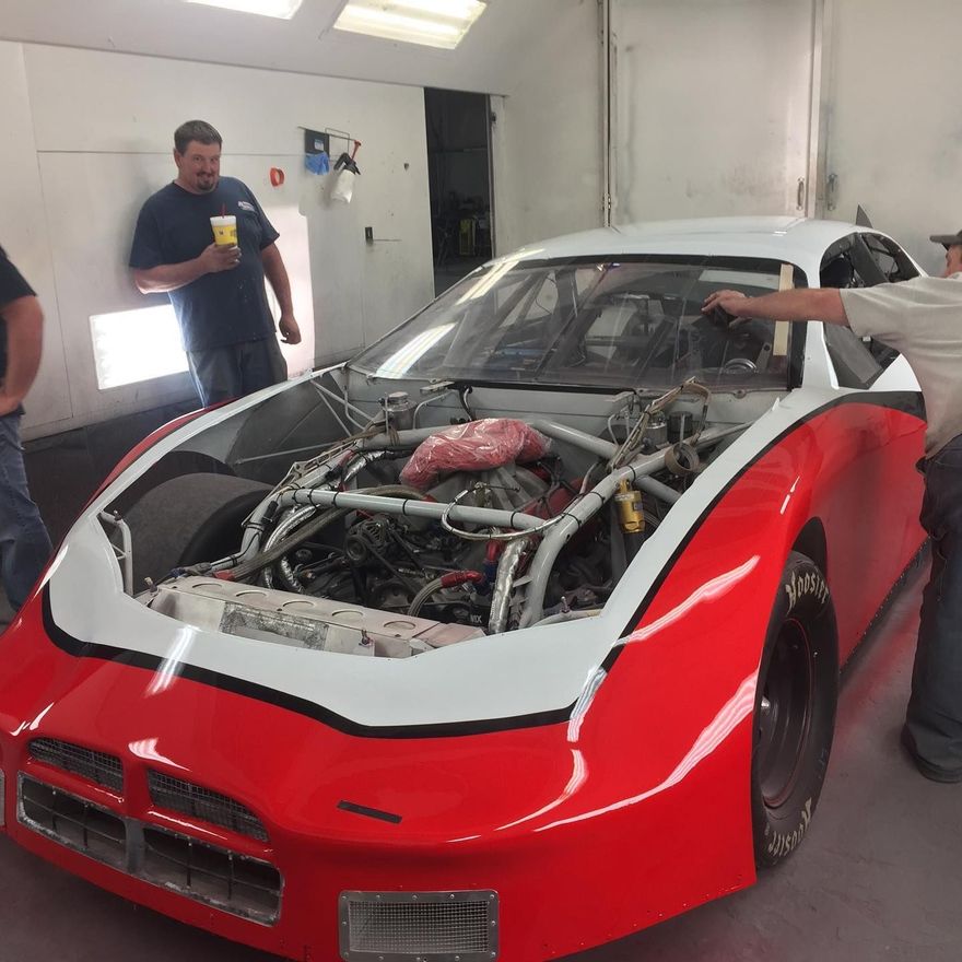 Red and white race car being worked on in a garage; two men look on.
