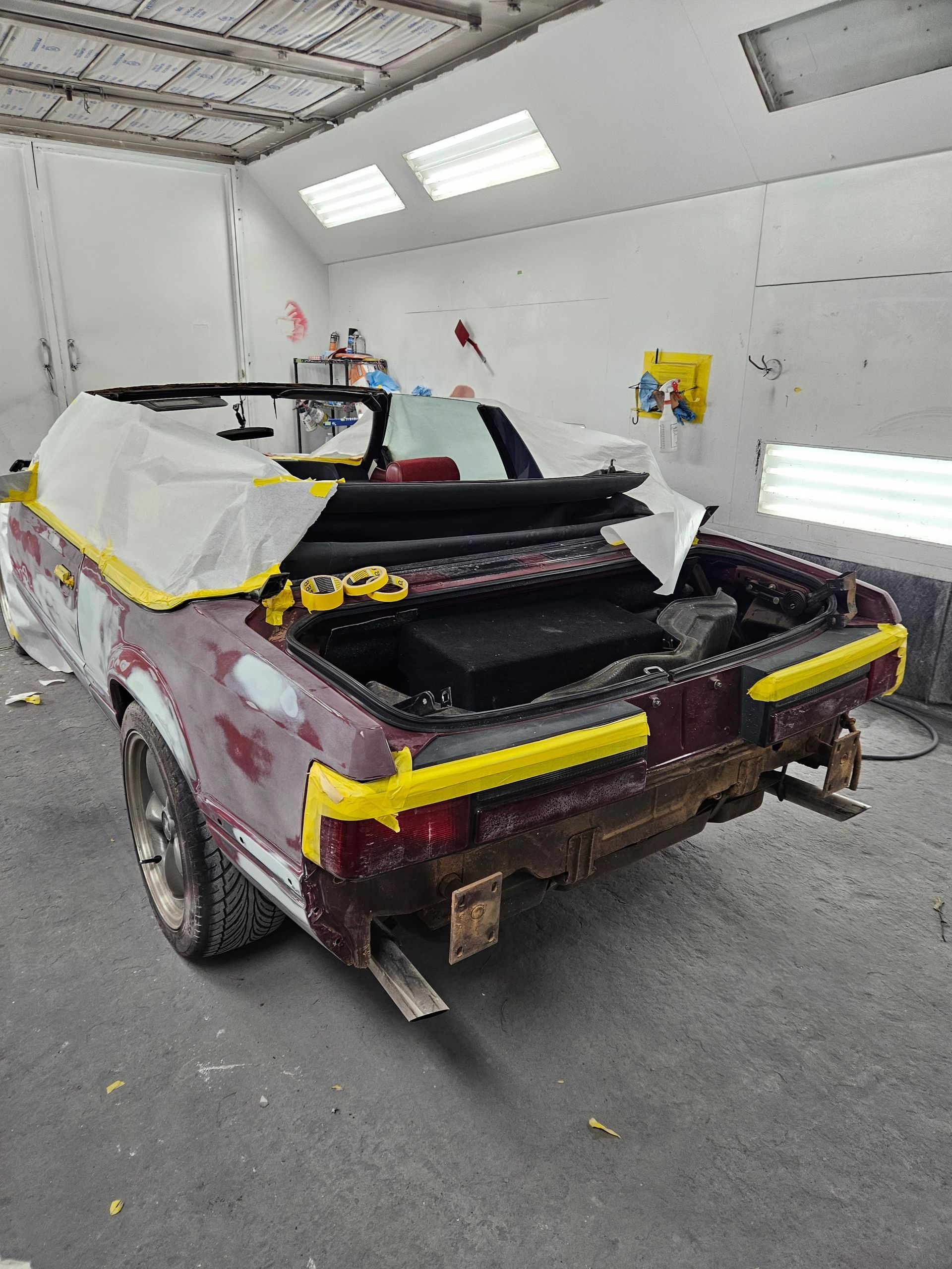 A convertible car in a paint booth, covered in tape. Rear view, maroon body, off-white roof.
