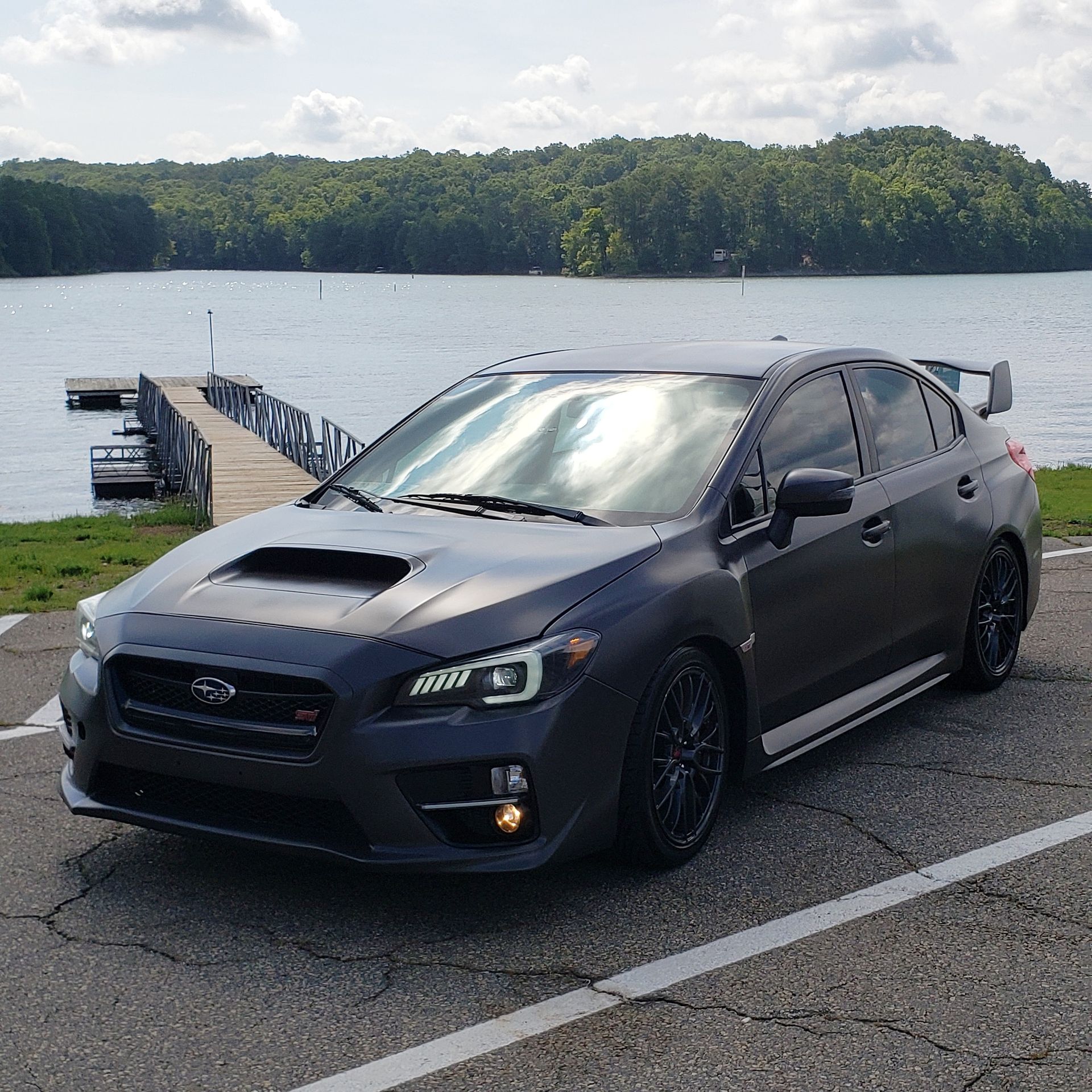 Dark gray Subaru WRX sedan parked by a lake, pier in the background, sunny day.
