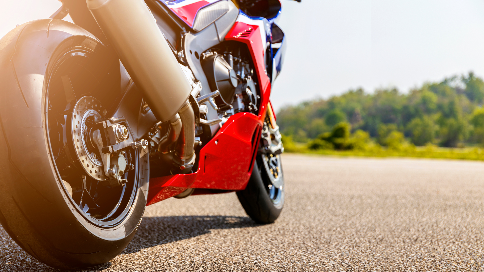 Red and blue sport motorcycle parked on asphalt, sunshine, trees in background.