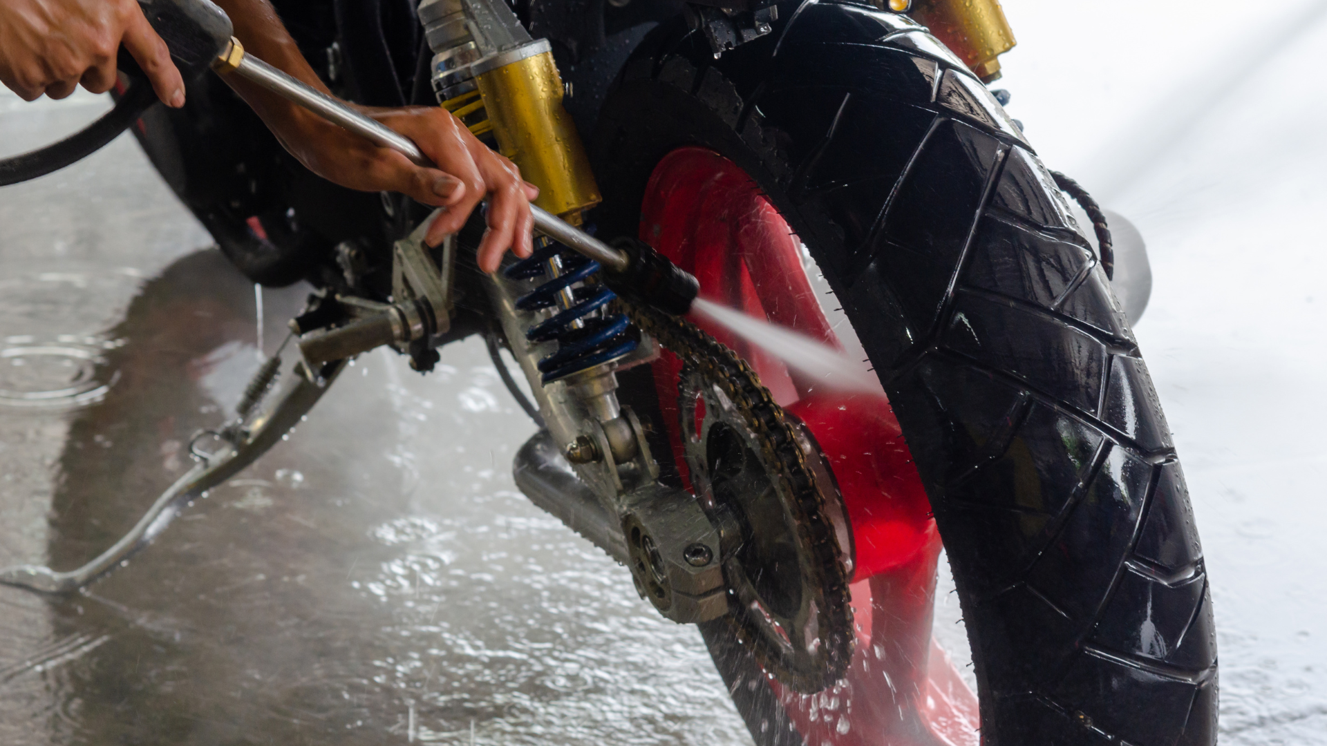 Motorcycle being washed with a pressure washer, focus on rear wheel and chain. Red wheel, black tire, hands holding hose.