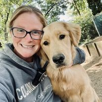 Woman smiling beside a golden retriever outdoors in a wooded area