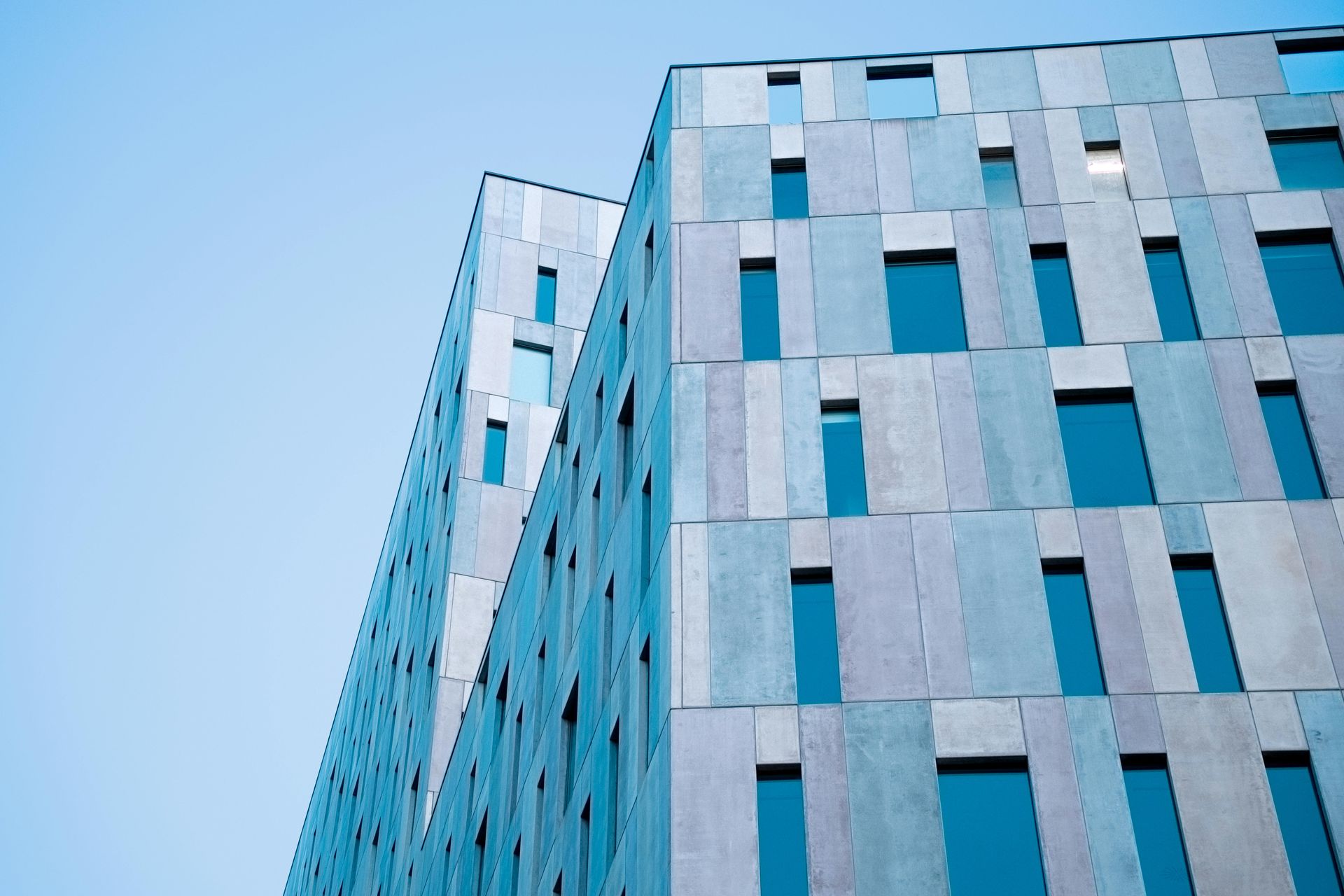 Modern building with a geometric facade of blue and gray panels and rectangular windows against a clear blue sky.