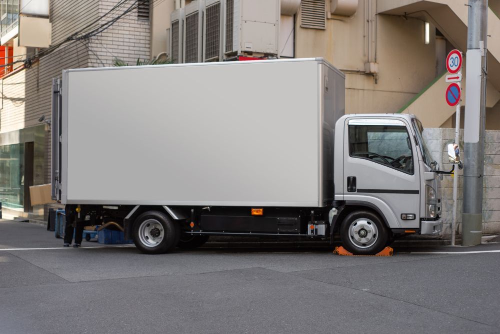 Delivery Truck Parked on a City Street, Silver Boxy Body, Beige Buildings — Hines Logistic Pty Ltd In Armidale, NSW