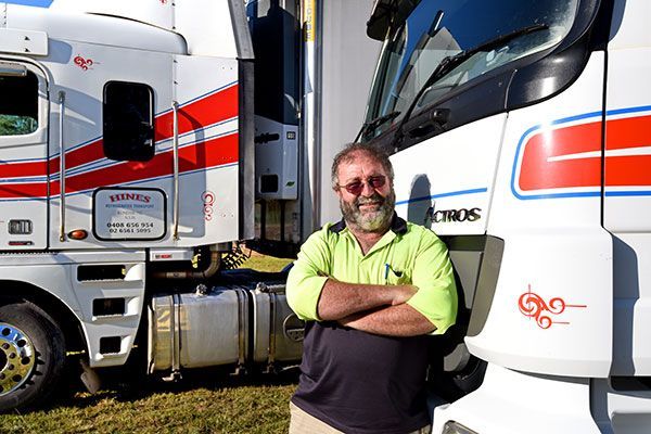 Man Standing With Arms Crossed, Smiling Next to Two White Trucks — Hines Logistic Pty Ltd In Kundabung, NSW