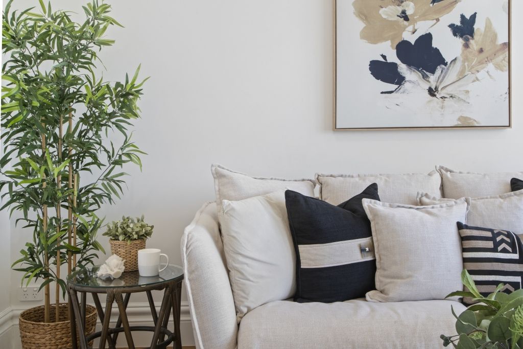 A living room with a white couch , pillows , a plant and a painting on the wall.