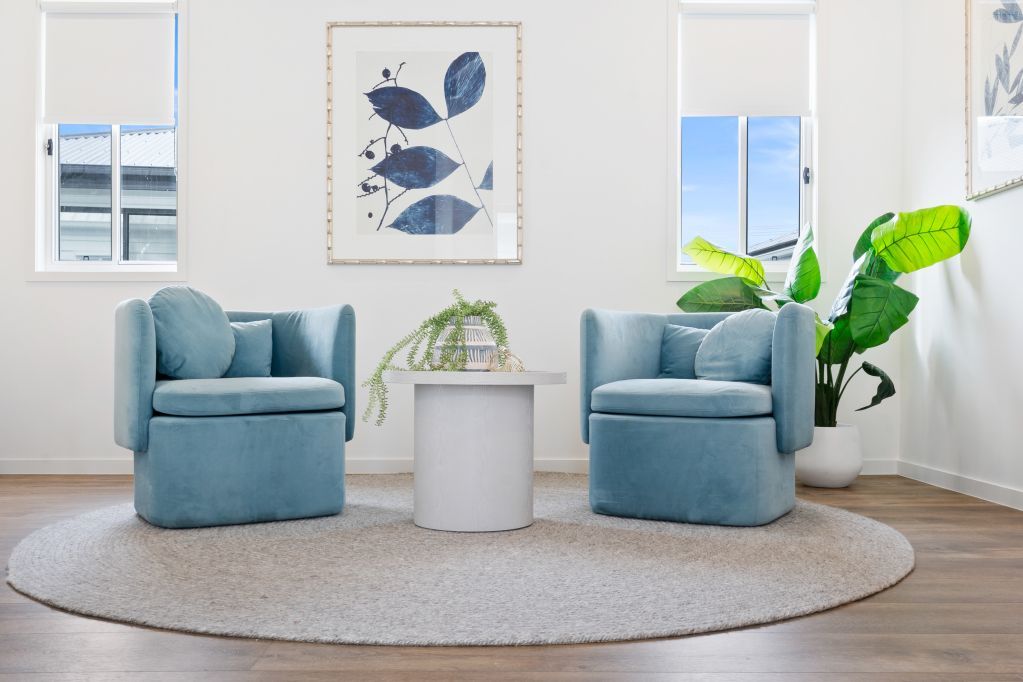 Bright living room with two blue chairs, round white table, large plant, and framed wall art on a rug.