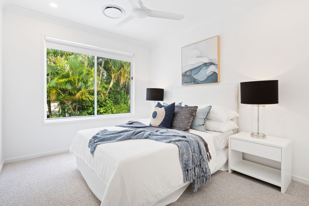 Bright white bedroom with a neatly made bed, nightstands, lamps, and a window with green foliage outside