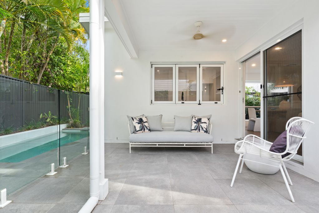 Covered patio with white seating, gray tiled floor, and a pool beside a black fence.