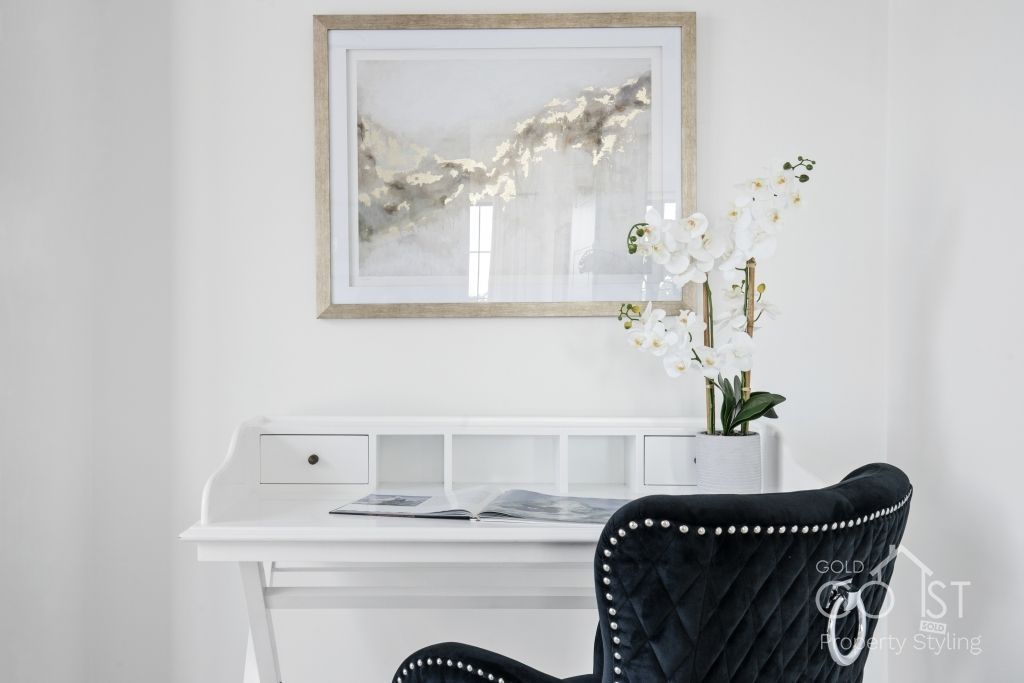 A black chair is sitting in front of a white desk with a picture on the wall above it.
