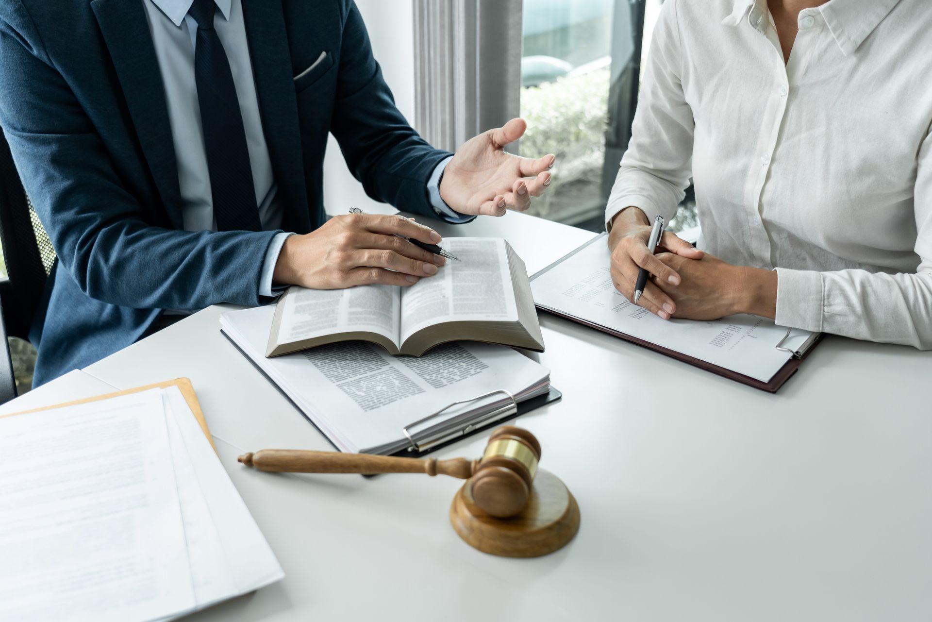 Lawyer in suit gesturing, consulting with a person at a table, legal documents, gavel, and book present.
