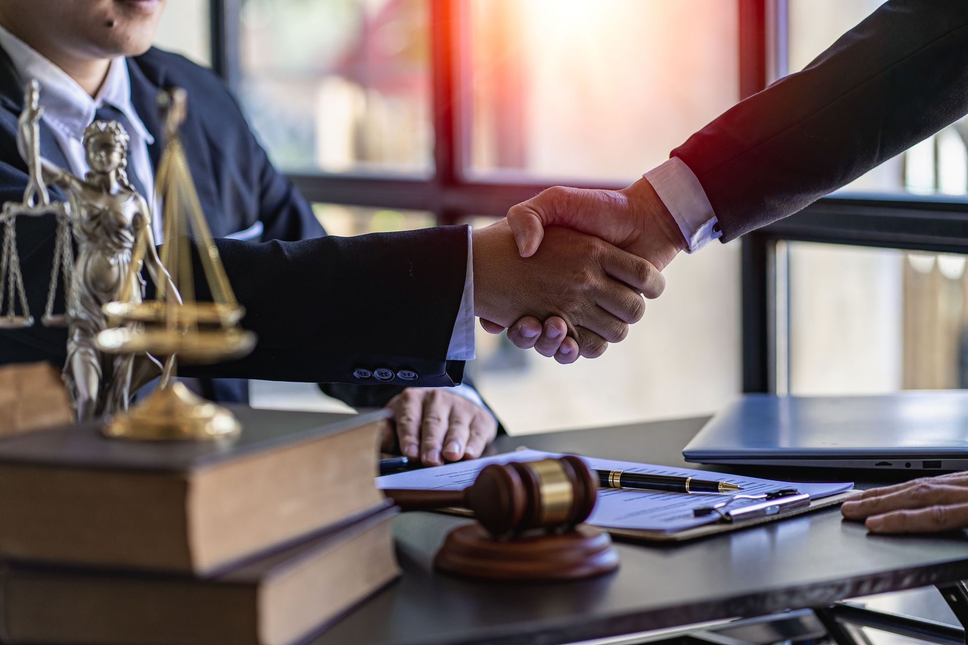 Lawyers shake hands at a desk with legal books, gavel, and scales of justice.