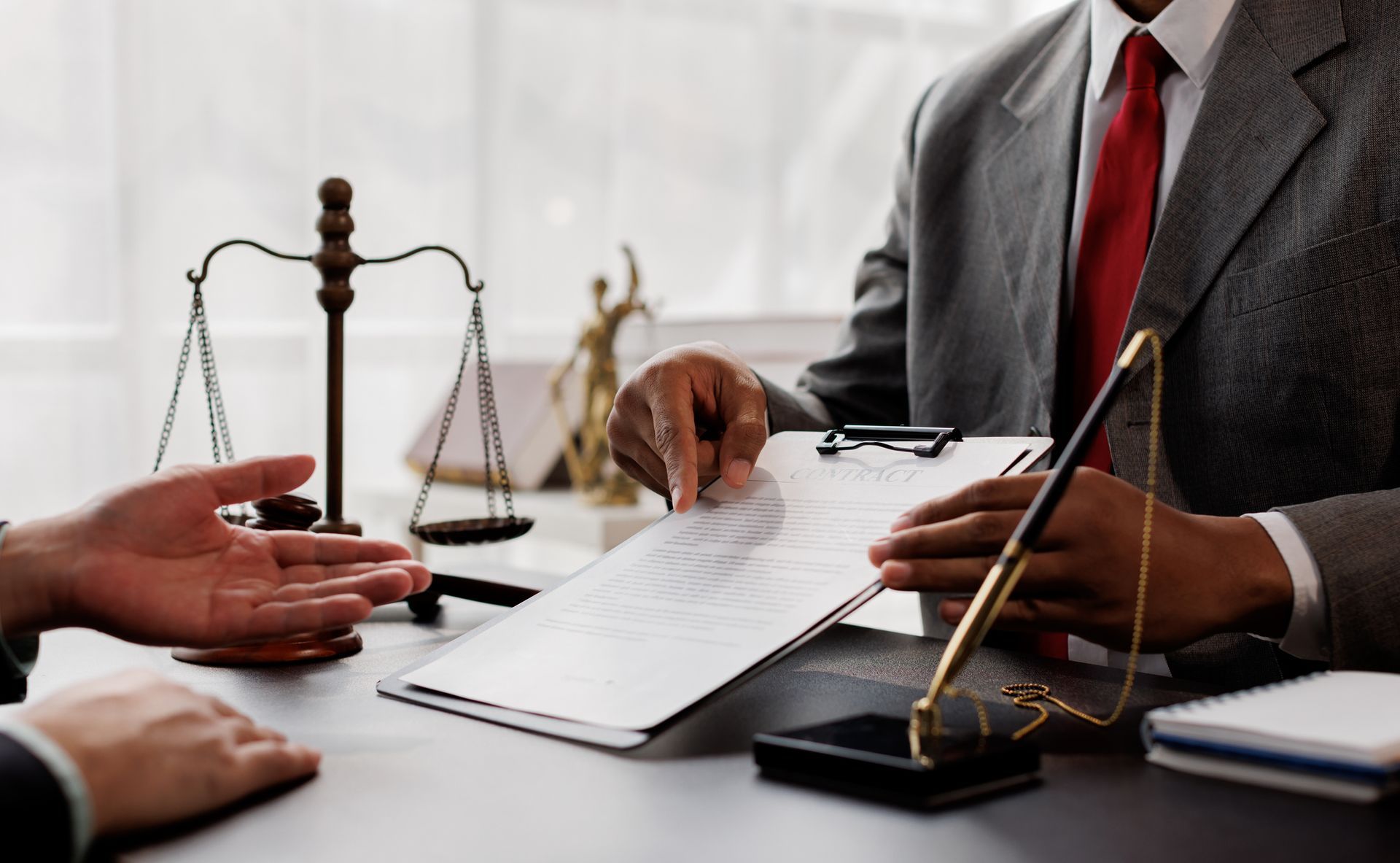 Lawyer in suit pointing to documents on a clipboard during a meeting with a client. Scales of justice in the background.