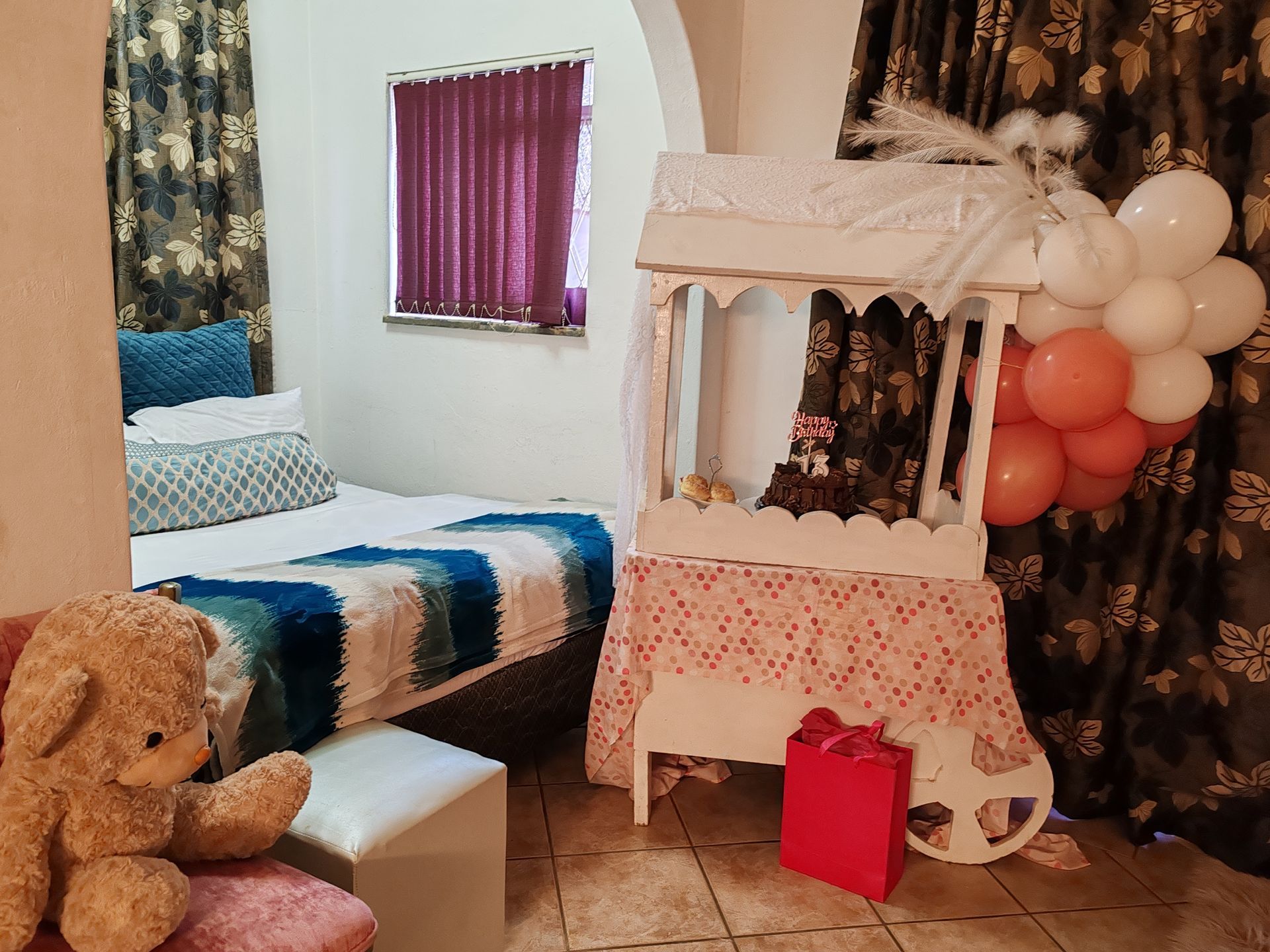 Decorated bedroom with bed, teddy bear, fairy lights, balloon cluster, and pink gift bag