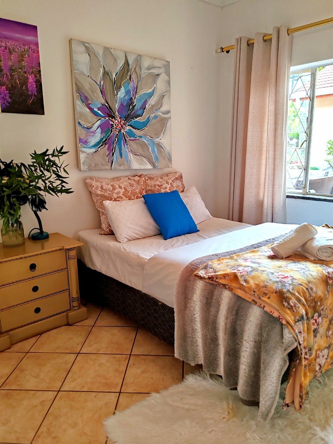 Cozy bedroom with a neatly made bed, colorful wall art, wooden dresser, potted plant, and sunlight by the window.
