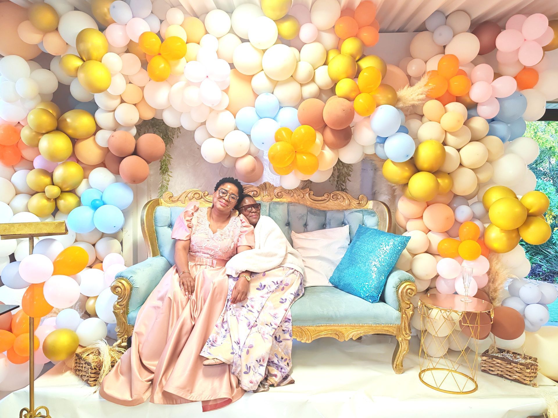 Two women sitting on a gold sofa in front of a pastel balloon arch backdrop