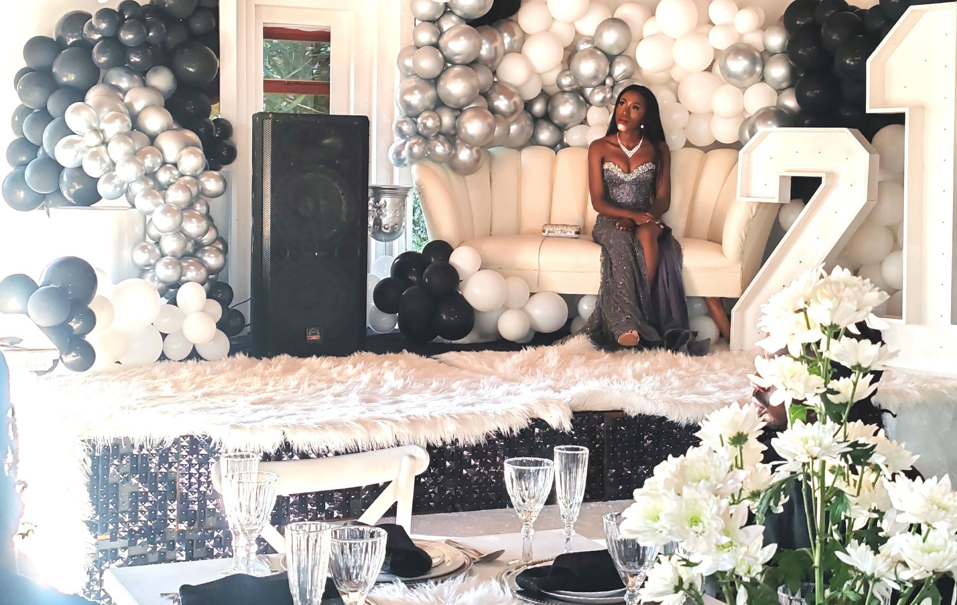 Decorated event stage with black, white and gold balloons, a large “7,” flowers, and a seated woman in a dark dress