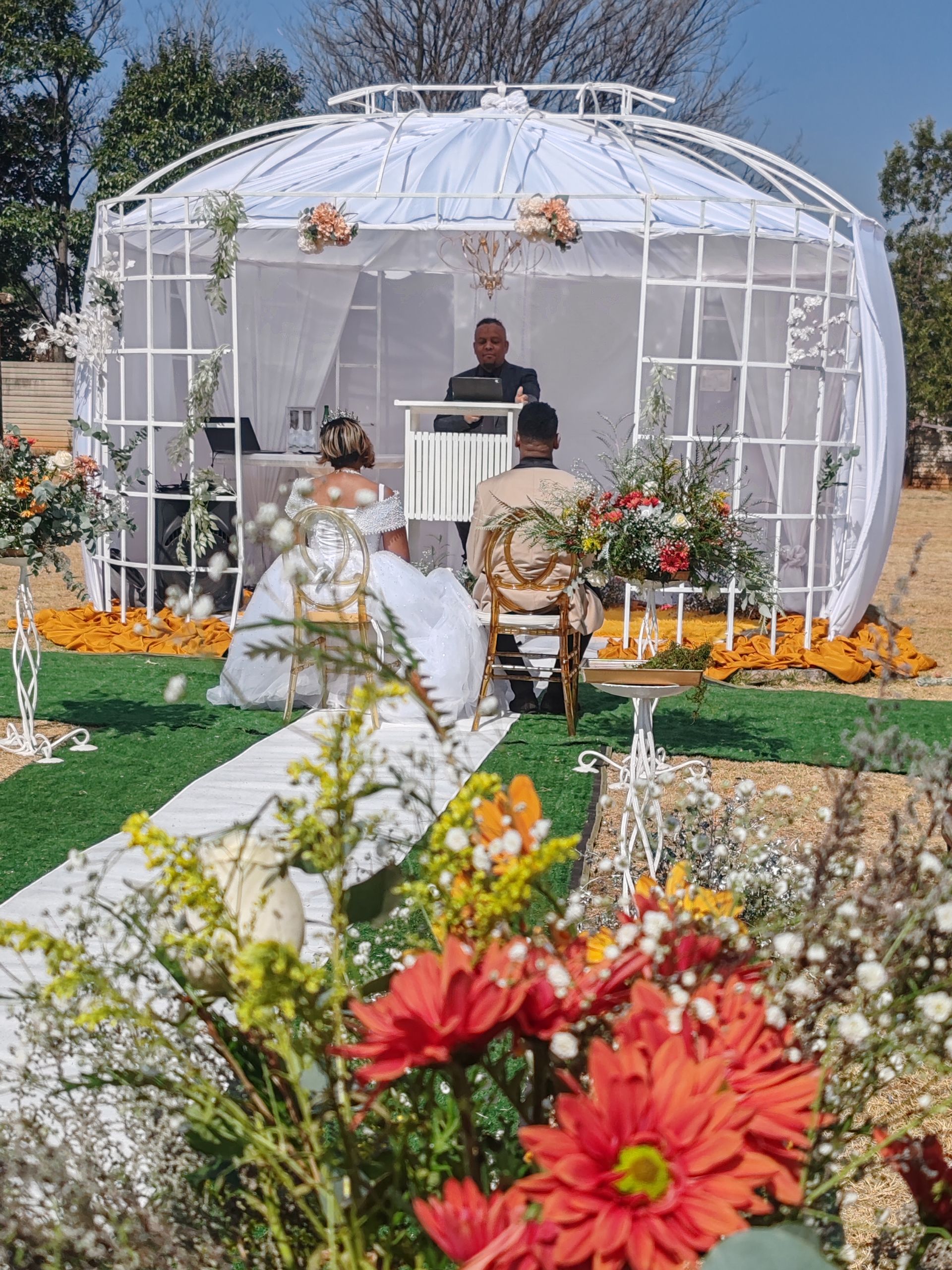White wedding gazebo decorated with flowers, with a person standing inside amid a garden ceremony.
