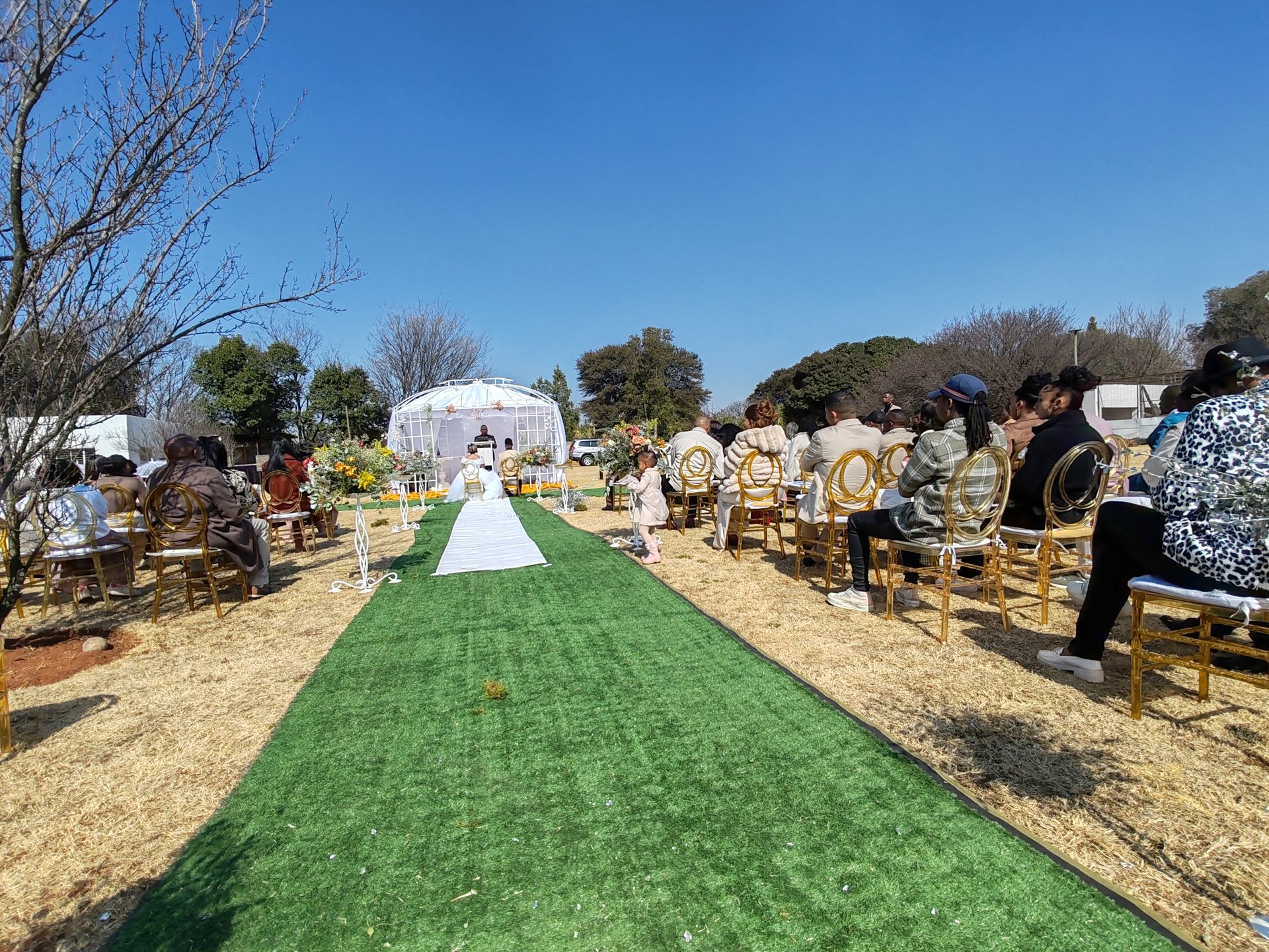 Outdoor wedding ceremony with guests seated on both sides of a green aisle under a clear blue sky