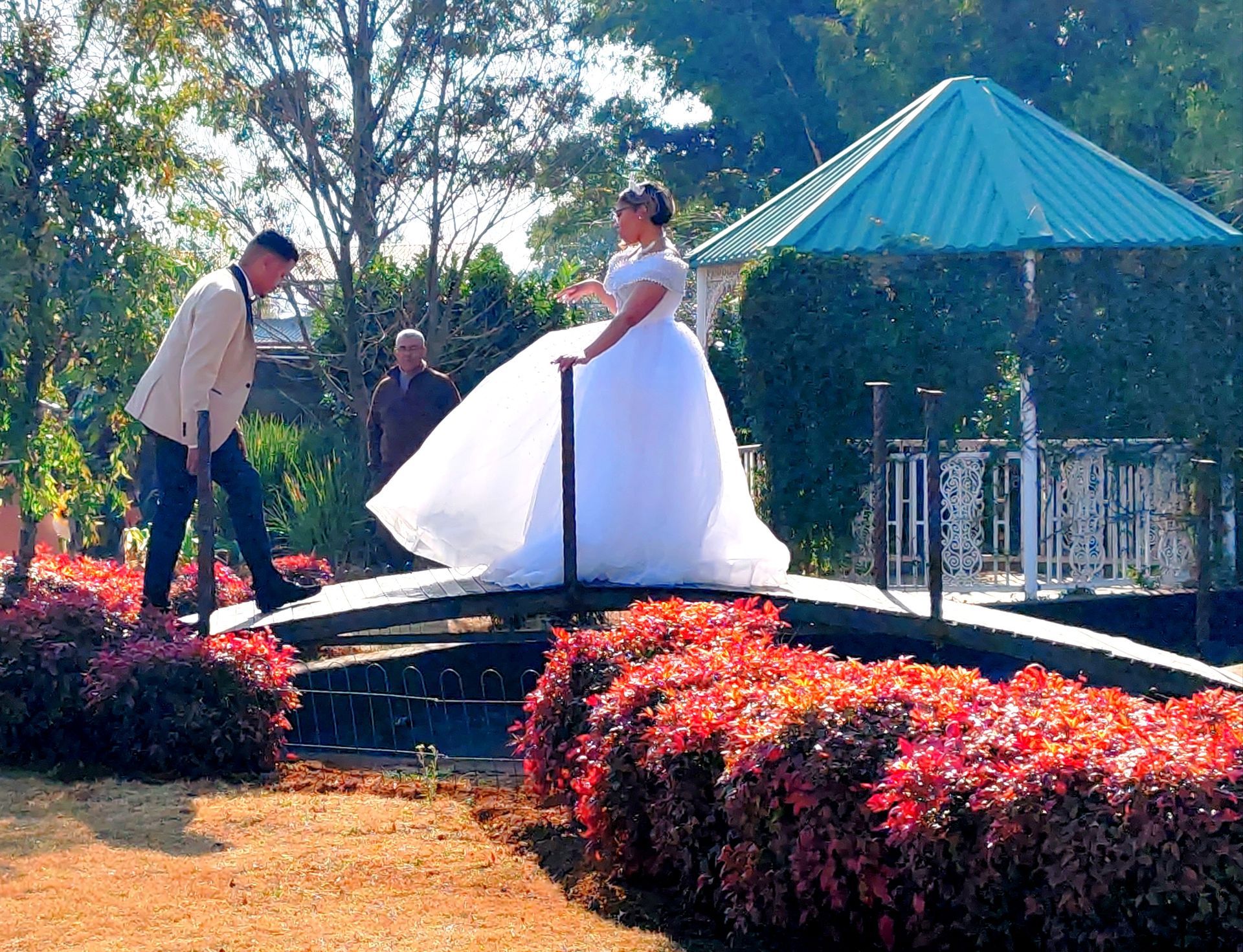 Bride and groom on a garden bridge, with a gazebo and red flowers behind them.