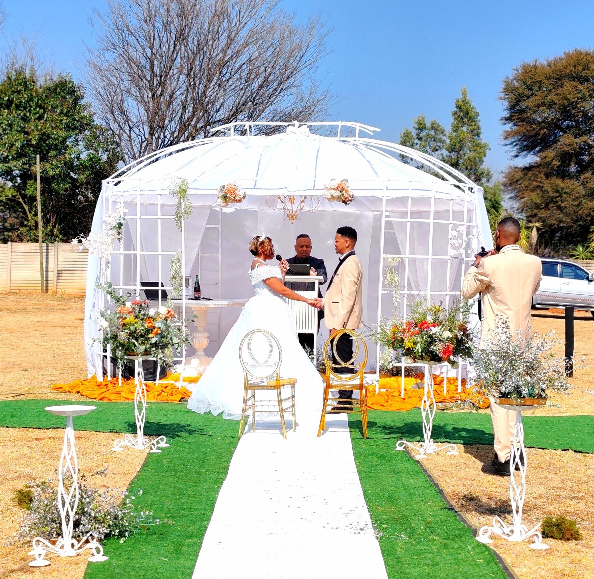 Wedding ceremony under a white gazebo with couple at the altar on a white aisle, floral decorations, and officiant