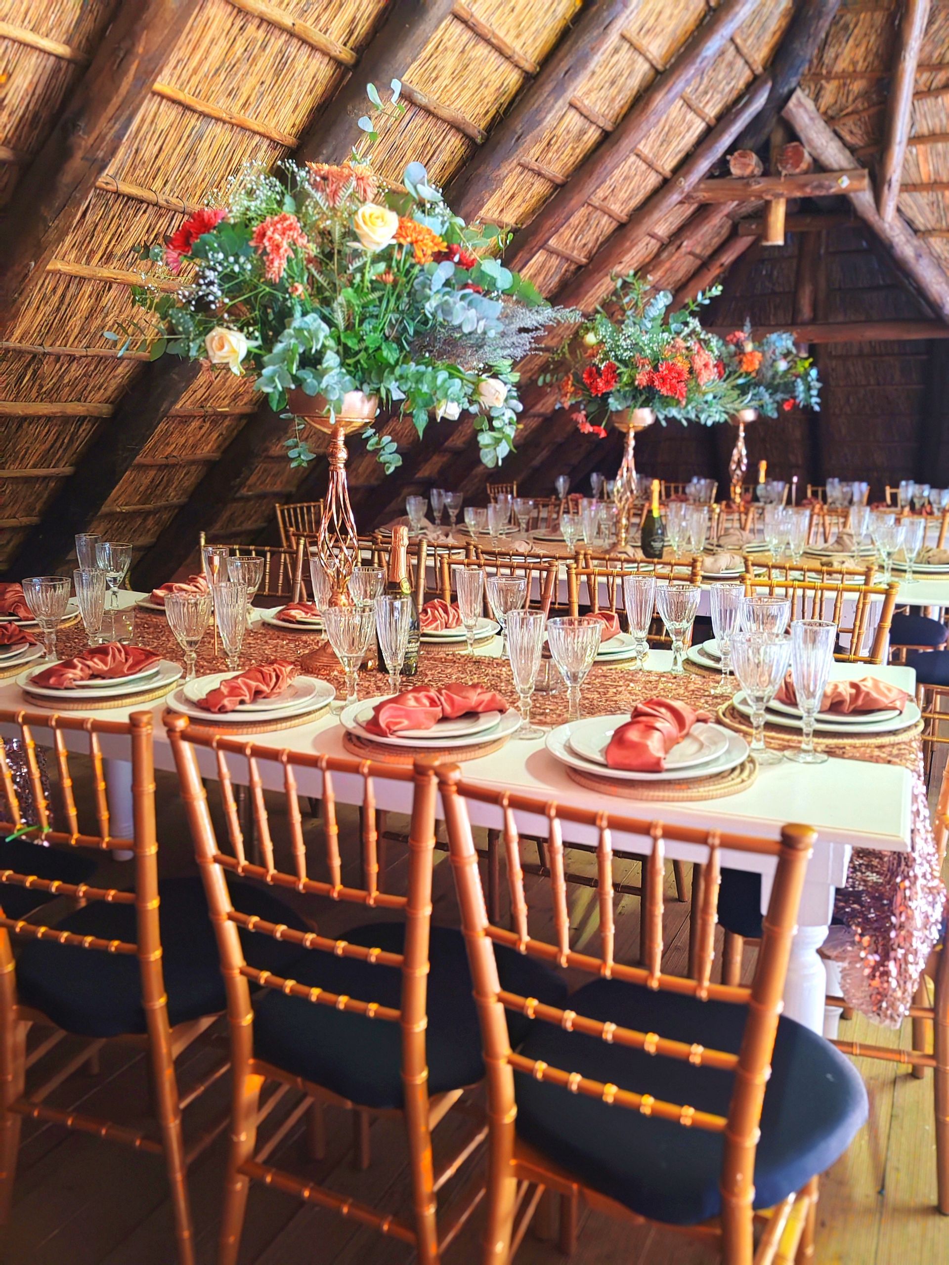 Banquet table set under a thatched roof with flowers, candles, and gold chairs