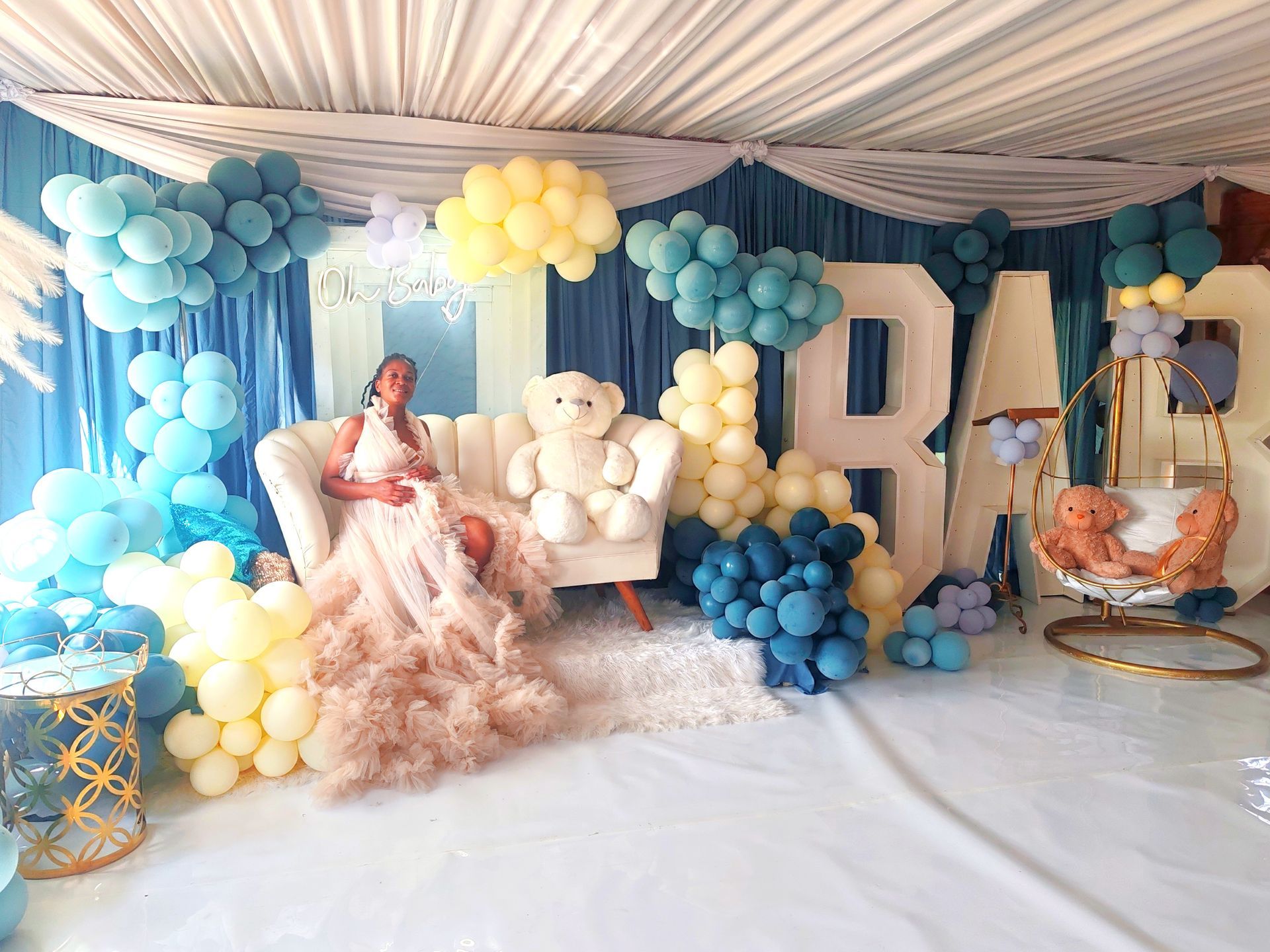 Blue and cream balloon-decorated celebration setup with teddy bears, large letters, and a seated woman in a pink gown