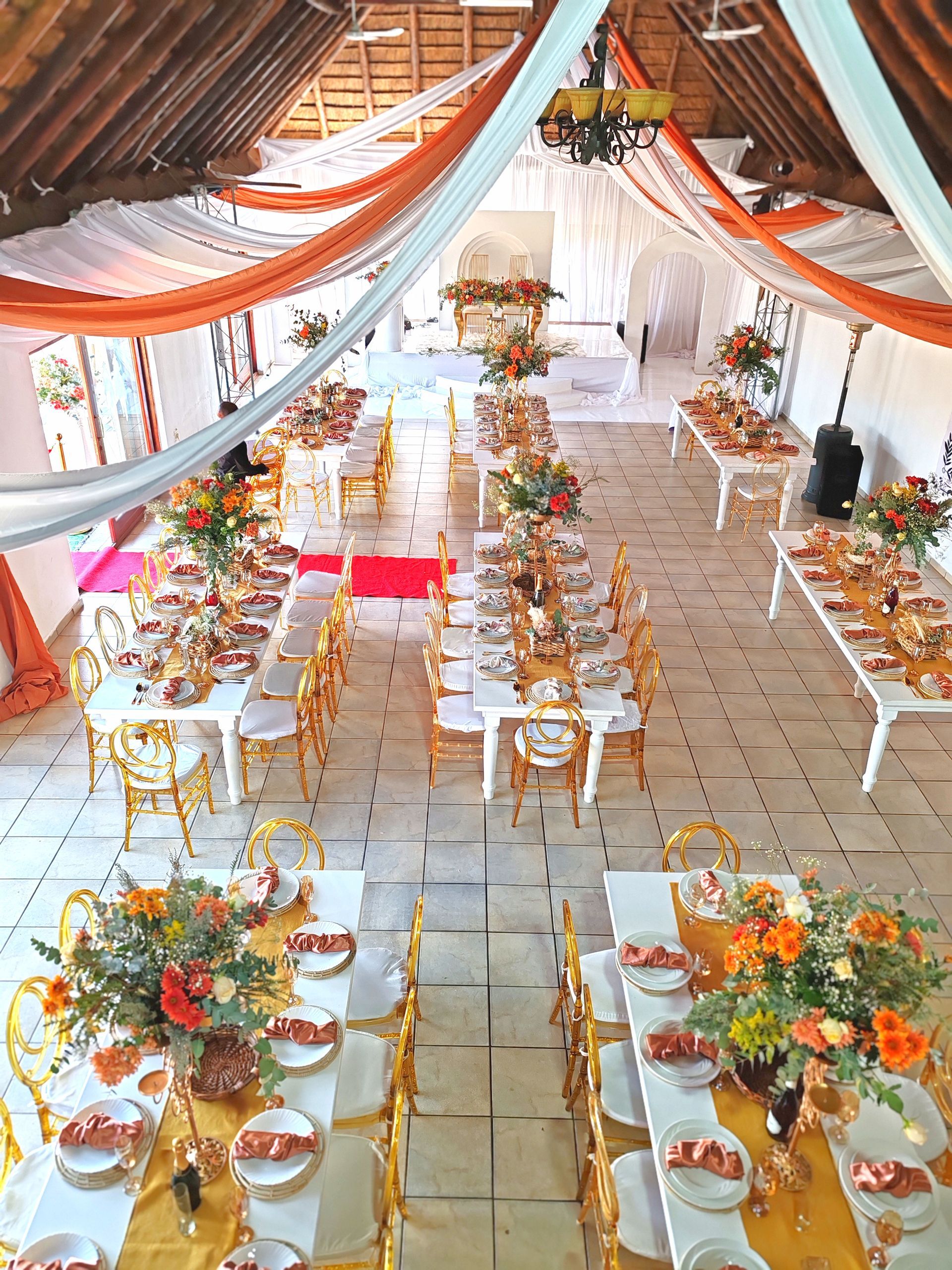 Decorated banquet hall with long tables, white chairs, and orange floral centerpieces under draped fabric