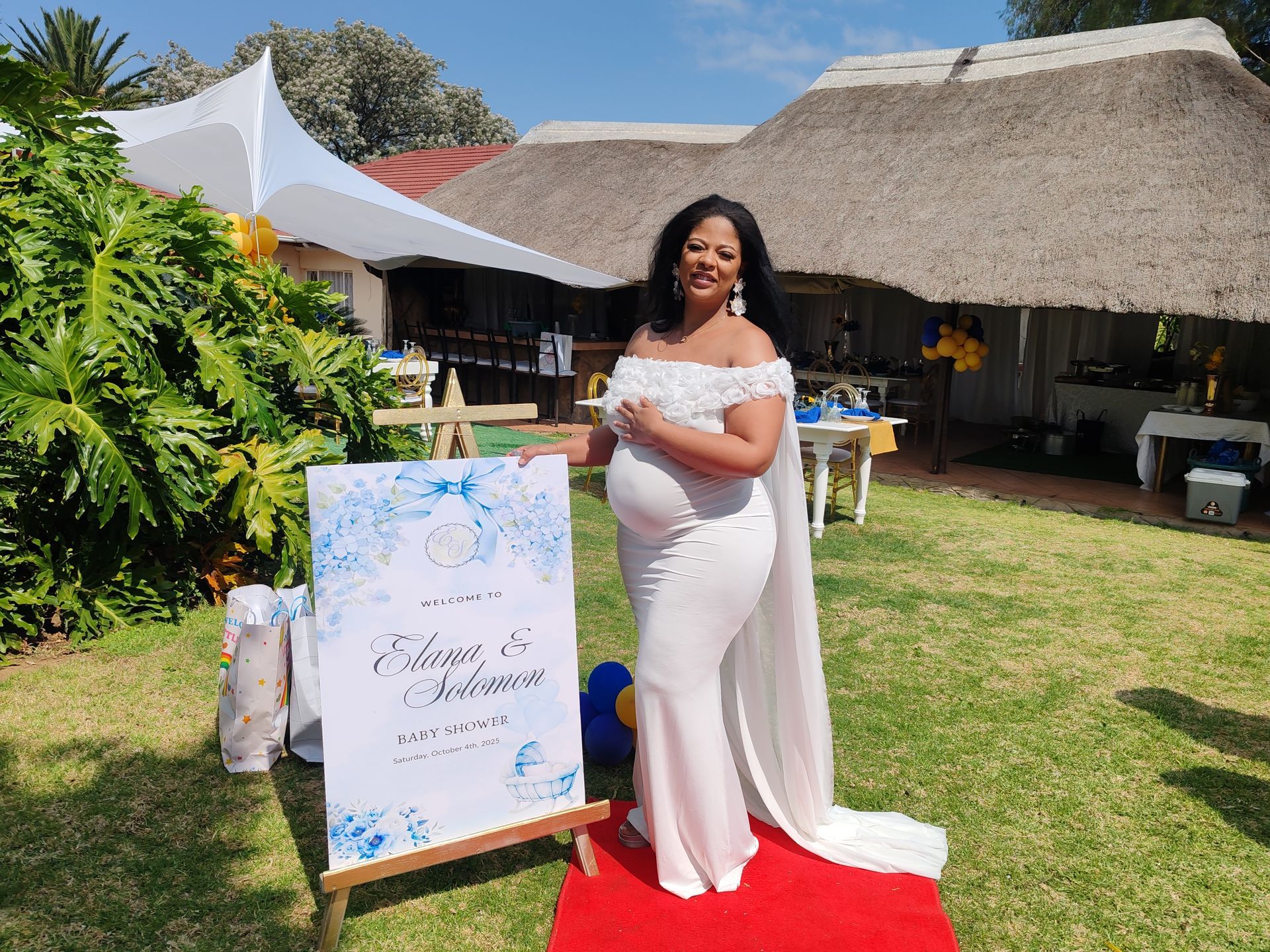 Pregnant woman in white dress posing beside a blue floral baby shower sign outdoors