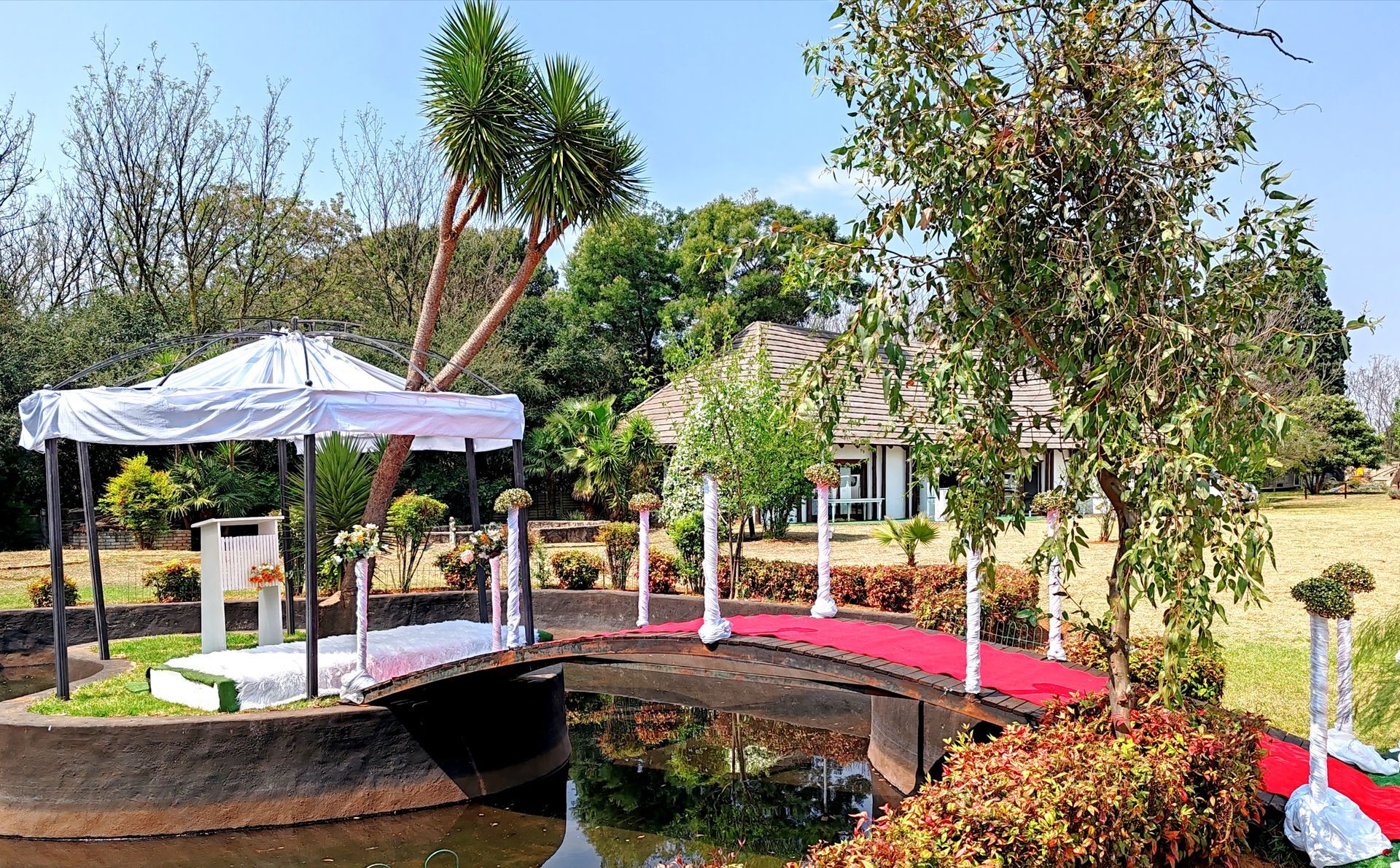 A red bridge over water in a lush garden with trees and white canopies on either side.