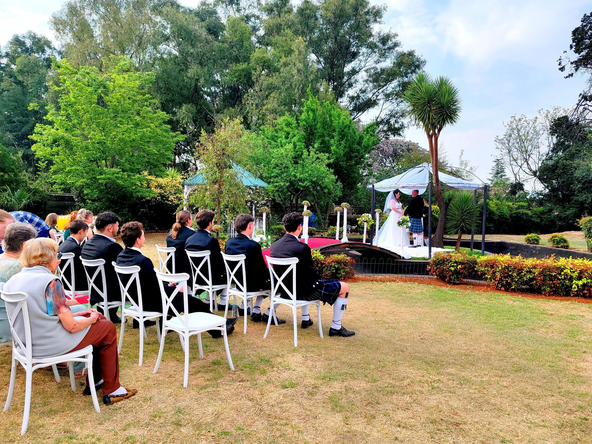 Outdoor wedding ceremony with guests seated on white chairs in a garden, facing an officiant under a floral arch.