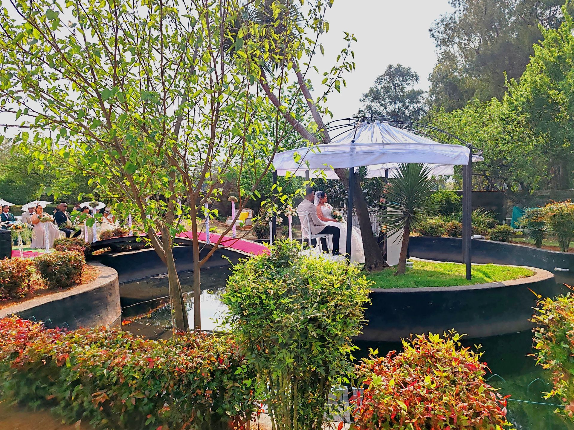 Lush garden patio with white umbrellas, curved ponds, and people seated among trees and flowers
