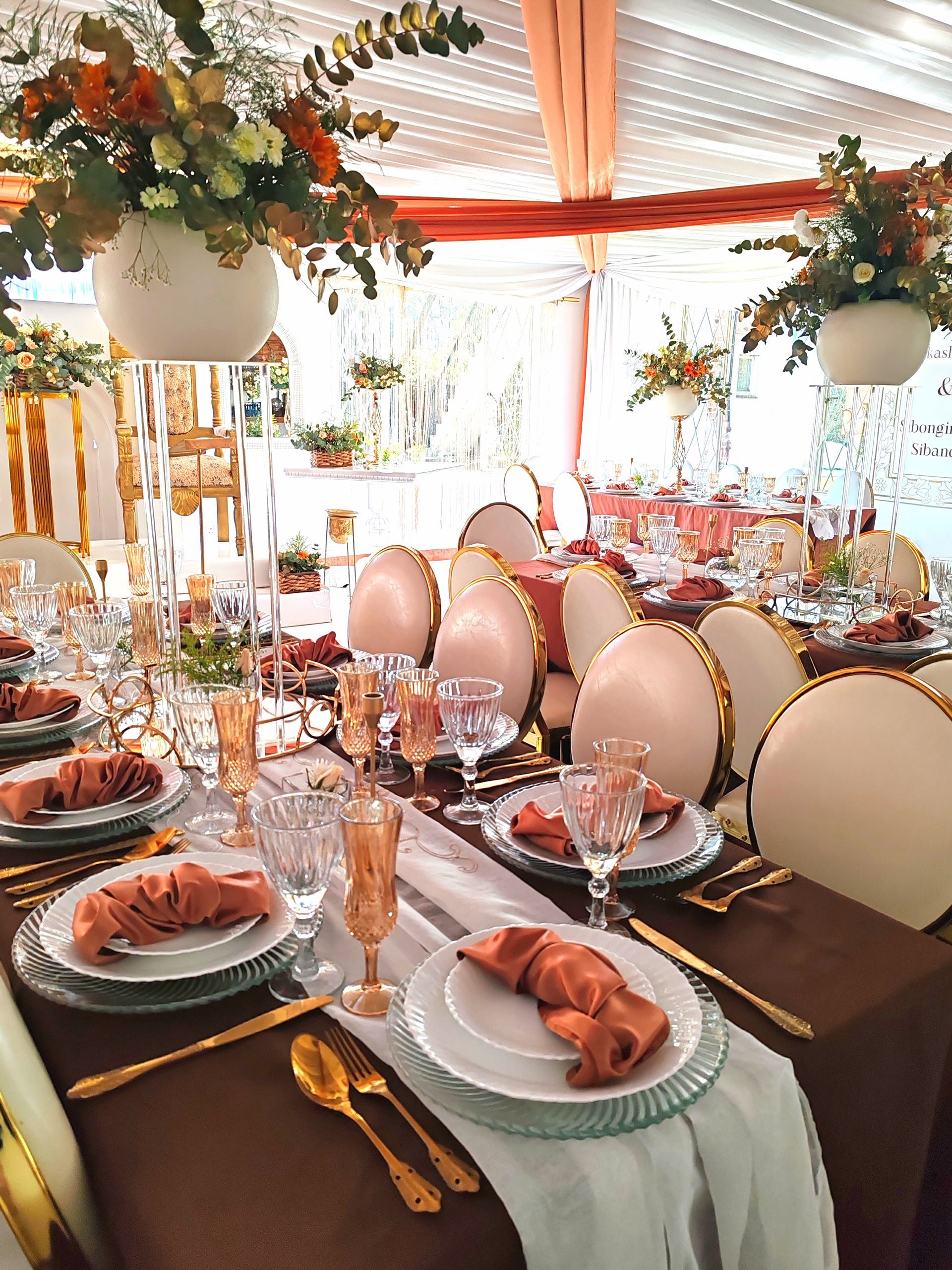 Elegant banquet table set with pink-and-gold chairs, floral centerpieces, and plates under a draped tent.