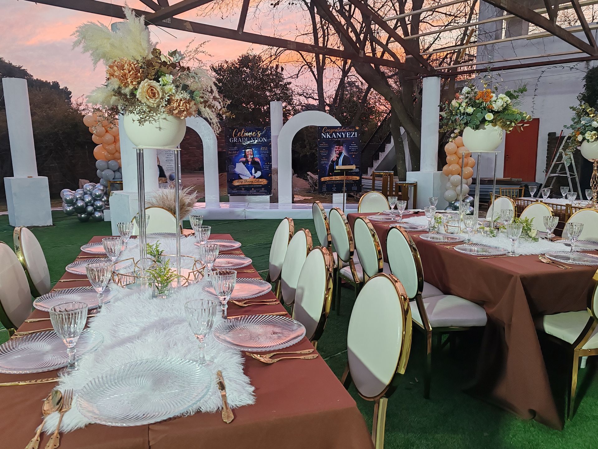 Elegant outdoor wedding reception setup with white chairs, floral centerpieces, and long tables under a tent at sunset