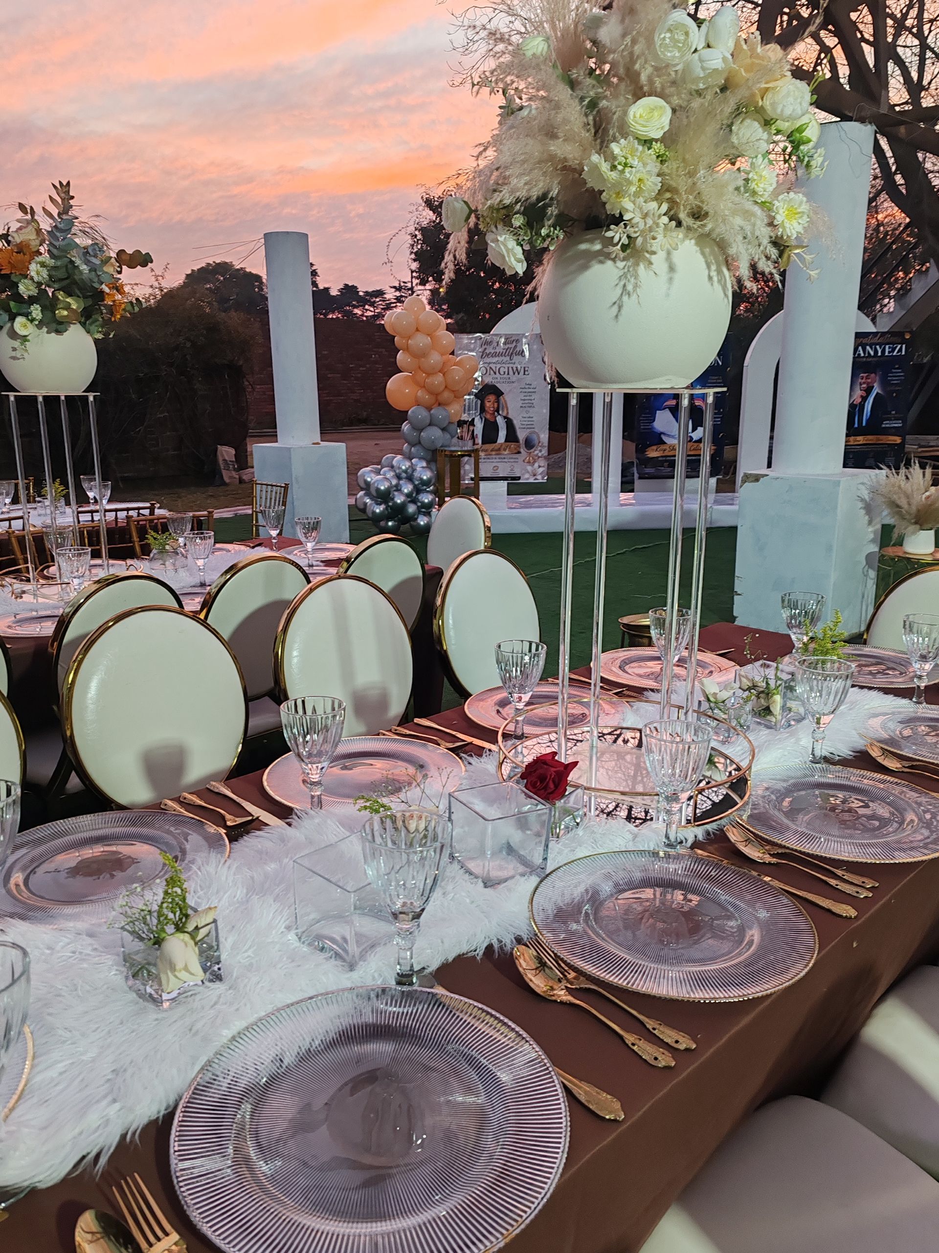 Elegant outdoor dinner table set with blush florals, clear glassware, and gold-trimmed plates at sunset