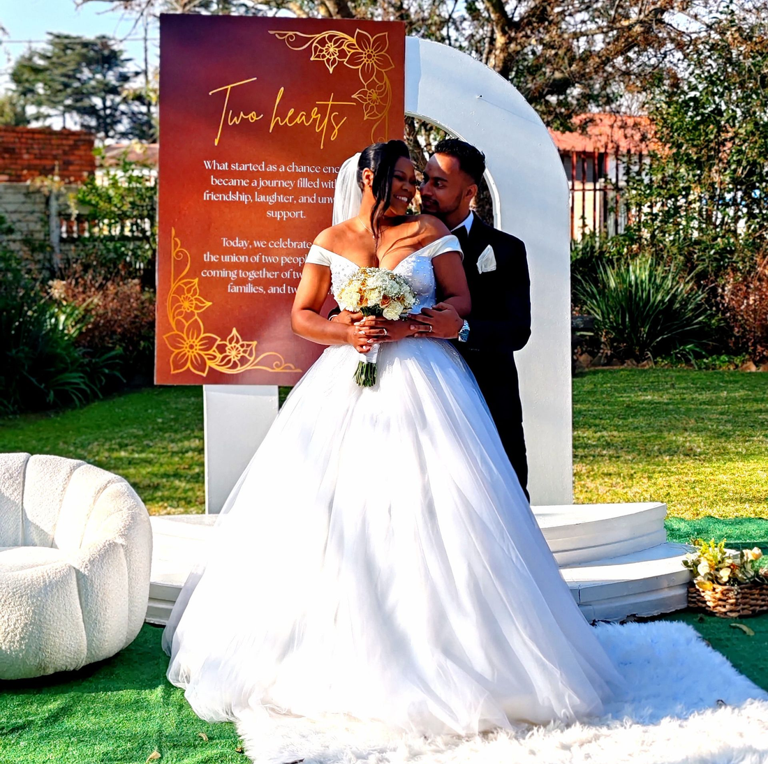 Bride and groom posing outdoors at a wedding ceremony with white floral decor and an orange backdrop