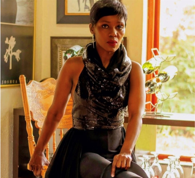 Woman in a black dress seated by a sunlit window, holding a chair in a warmly lit room