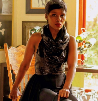 Woman in a black dress seated by a sunlit window, holding a chair in a warmly lit room