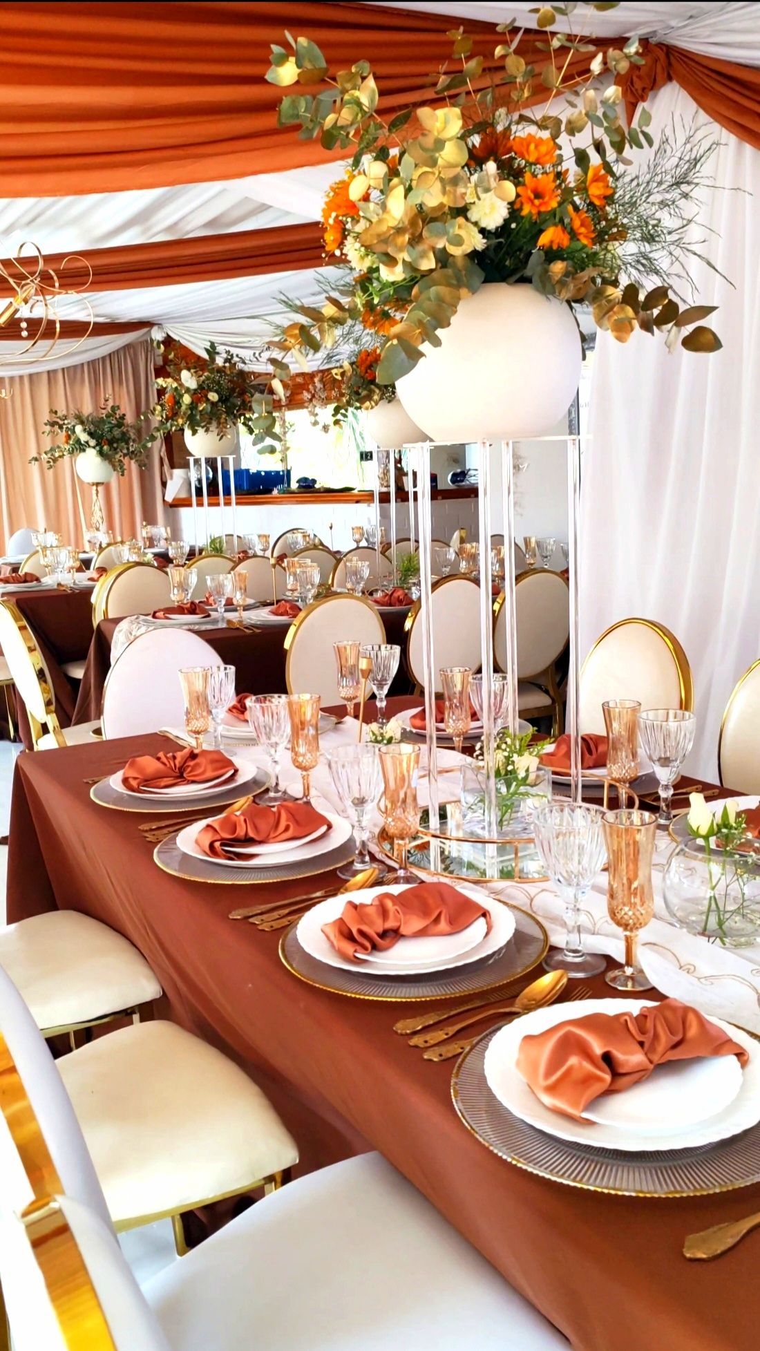 Elegant banquet table with orange napkins, white chairs, and floral decor under draped orange-and-white fabric.