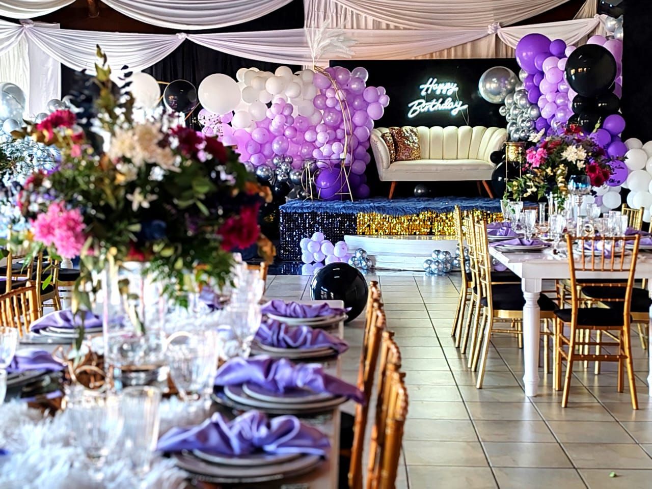 Decorated banquet hall with purple balloons, draped tables, and a stage at the far end