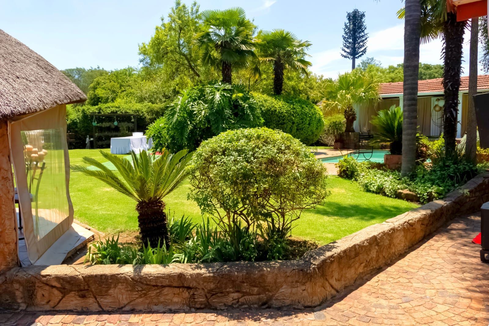 Lush landscaped garden with green shrubs, palm trees, and a stone path beside a thatched patio.