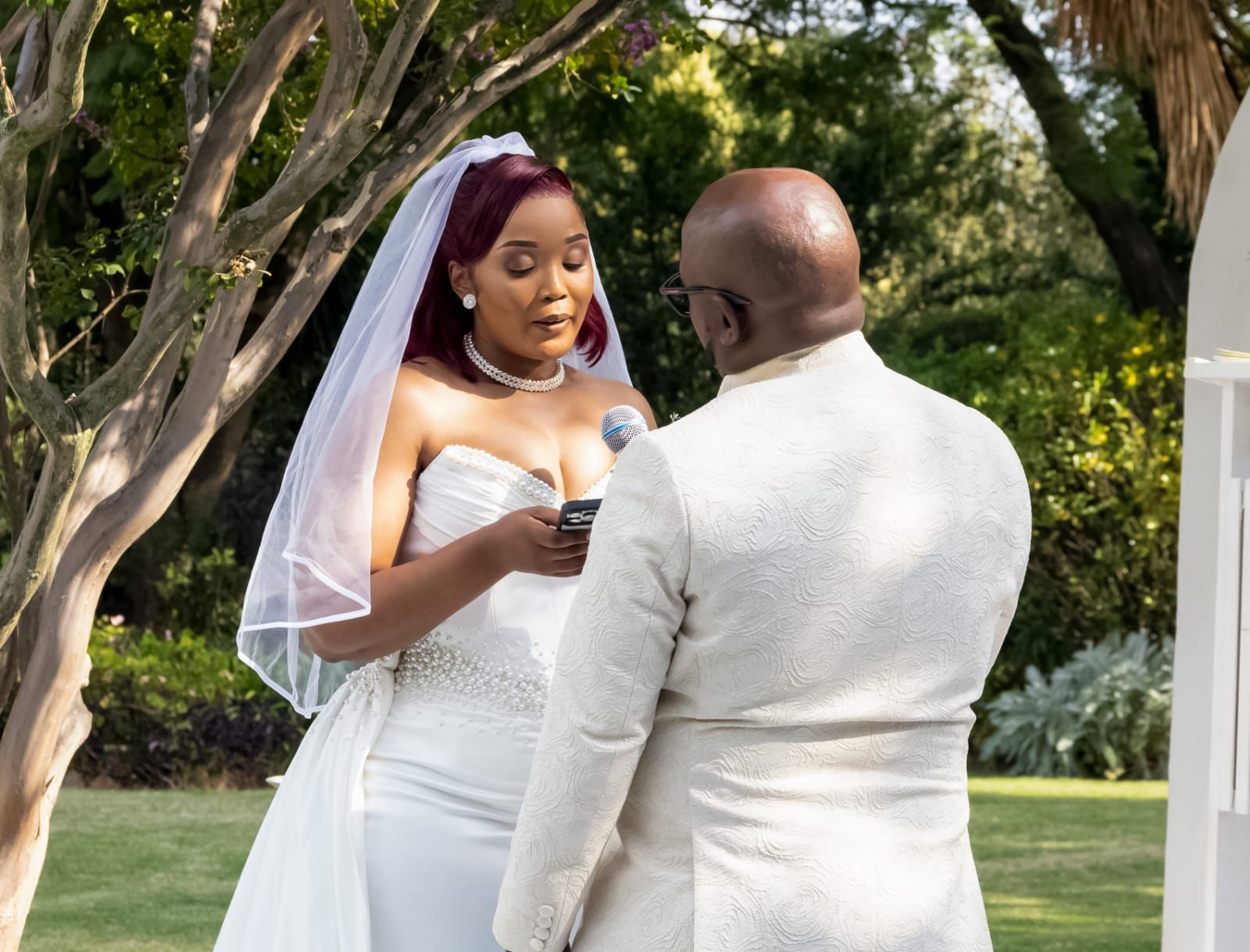 Bride and groom exchanging vows outdoors under trees, the bride holding a ring box.