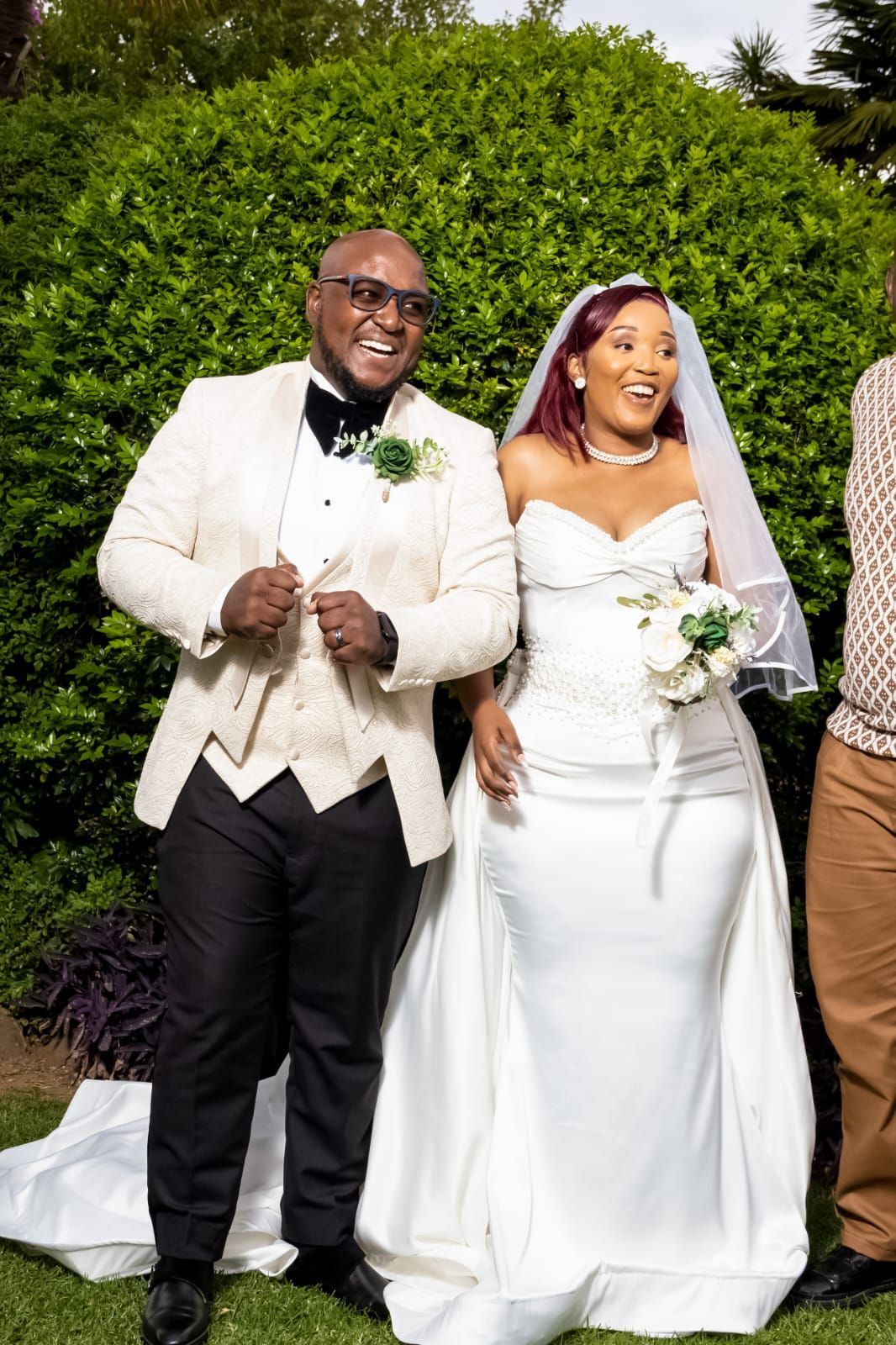 Wedding couple in white attire smiling outdoors in front of green bushes