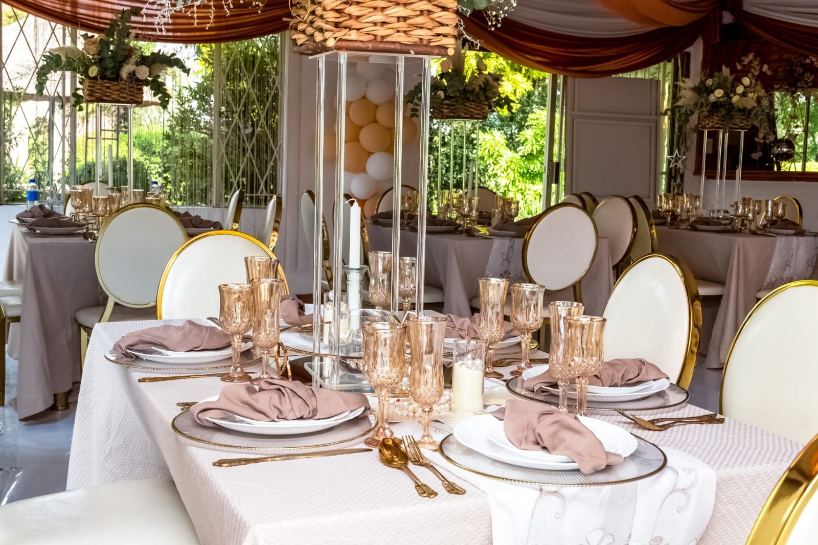 Elegant outdoor dining table set with white chairs, gold accents, and floral centerpieces under a canopy