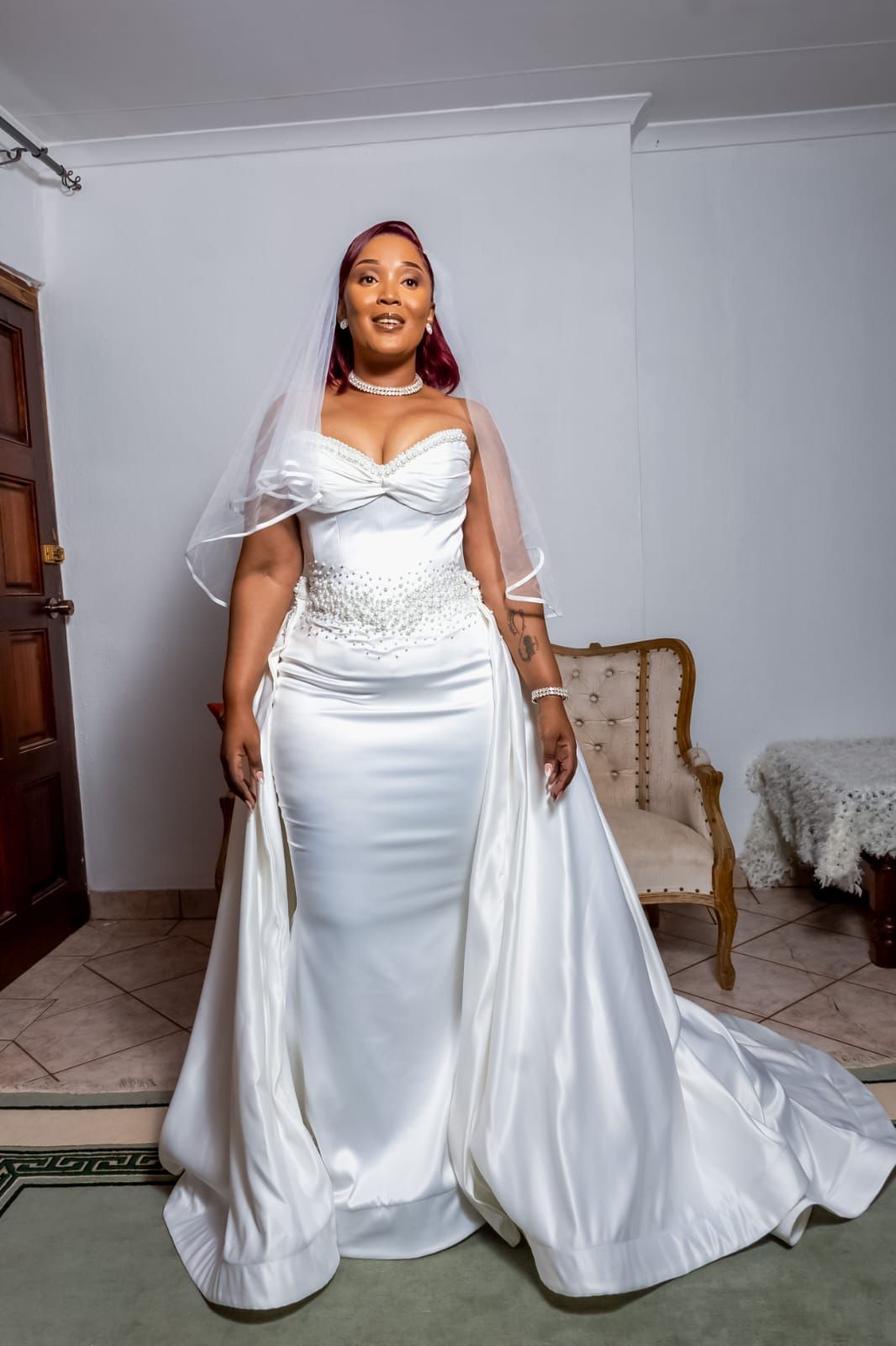 Woman in a white satin wedding gown and veil standing indoors, smiling beside a chair.