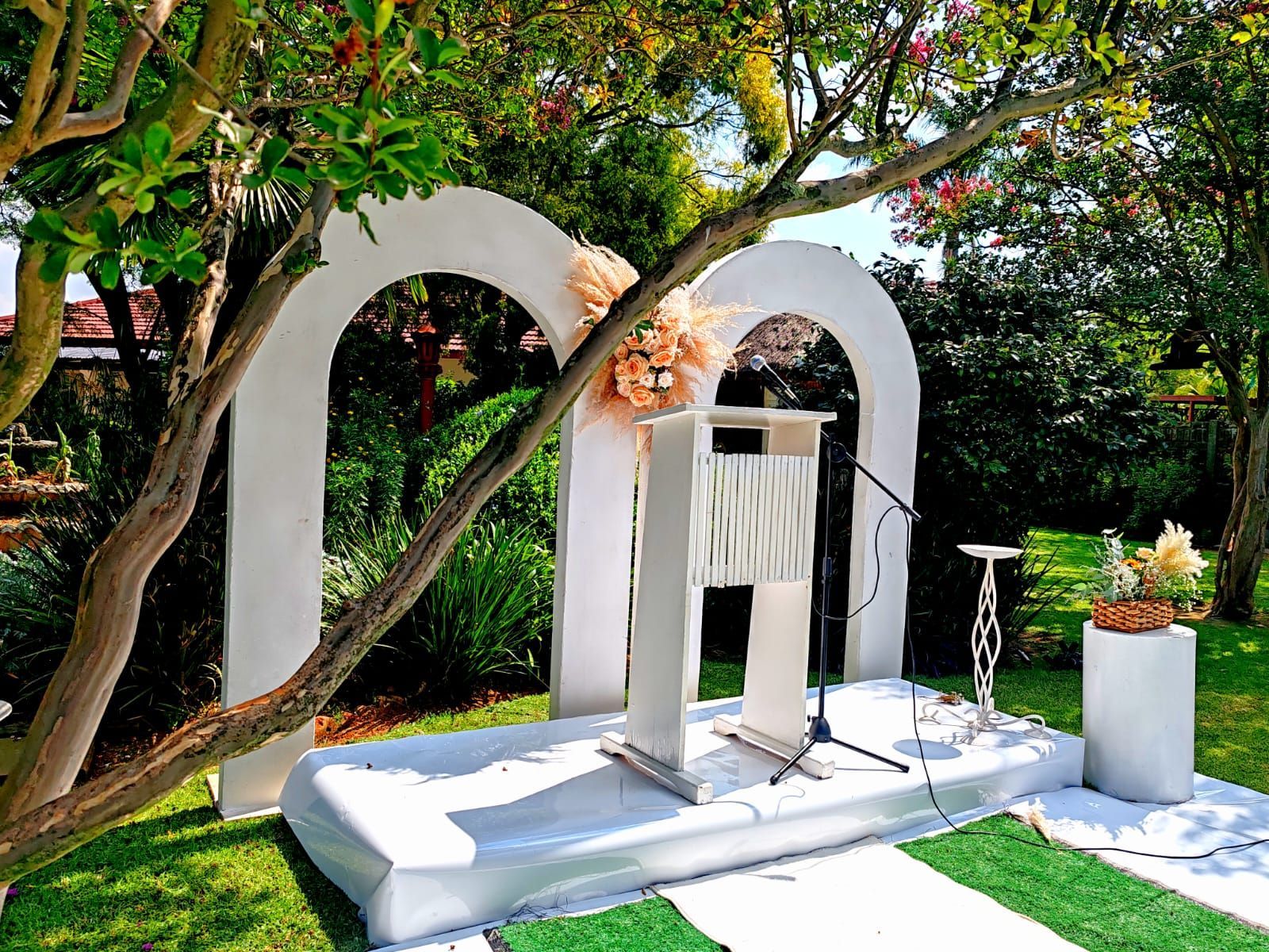 Outdoor white archway altar with floral decor, white benches, and greenery in a garden setting