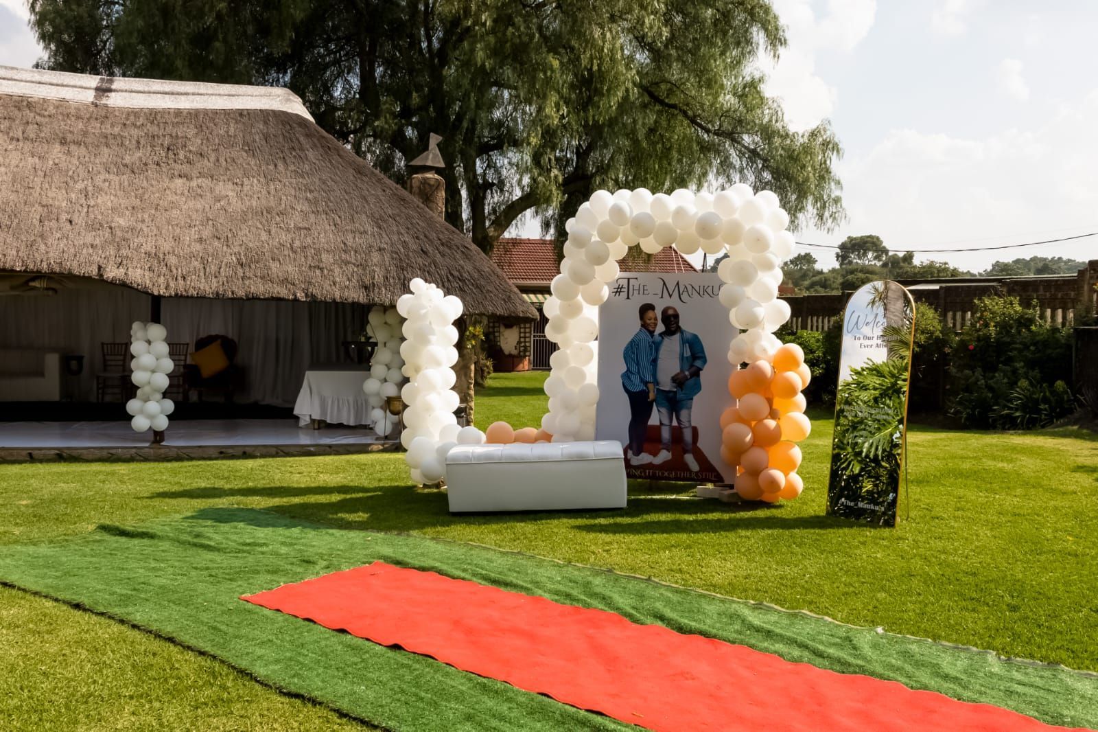 Outdoor event setup with white and orange balloon arch, red carpet, and people posing on a lawn