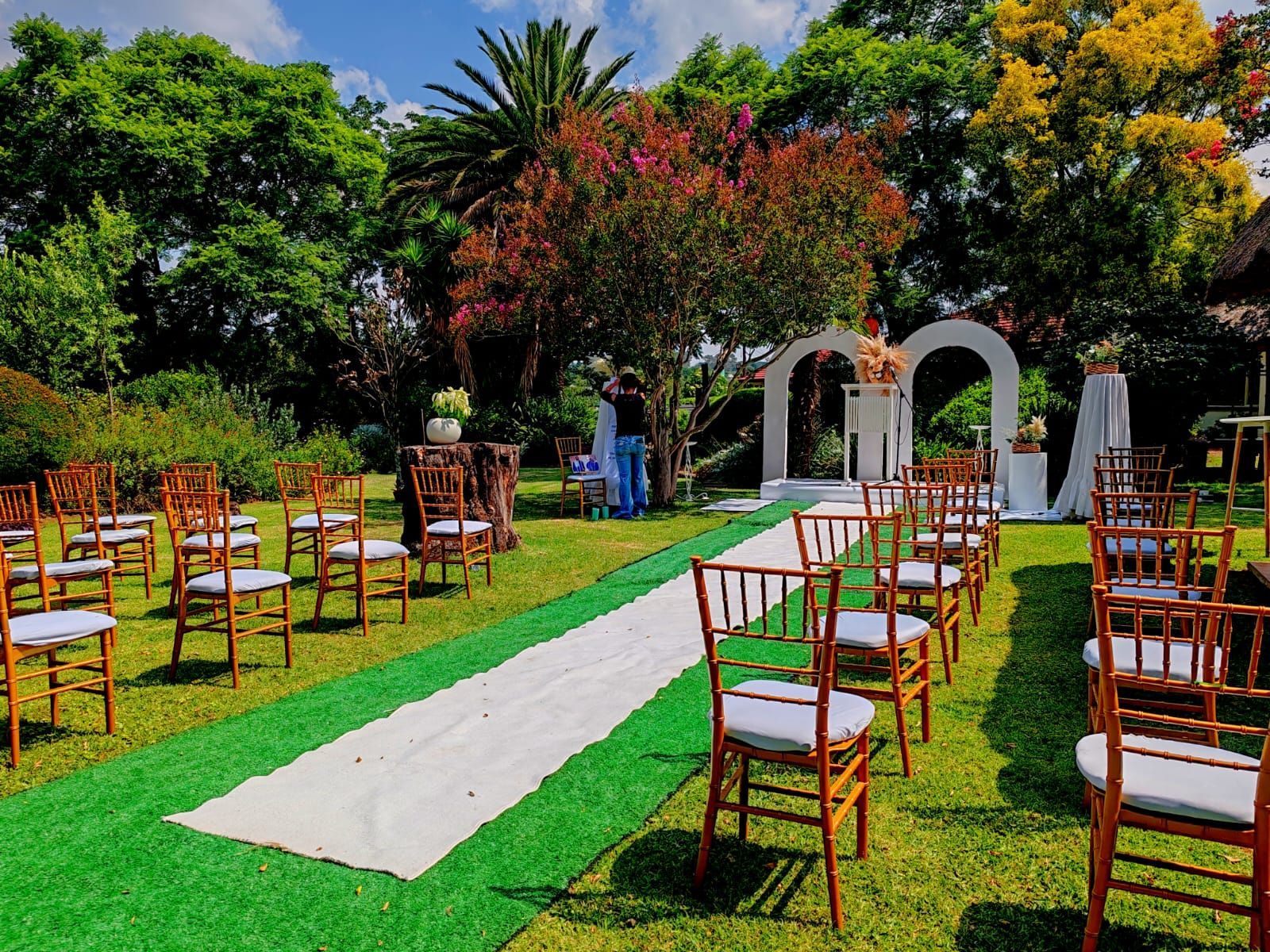 Outdoor wedding aisle with wooden chairs on a green lawn, bordered by trees and colorful flowers