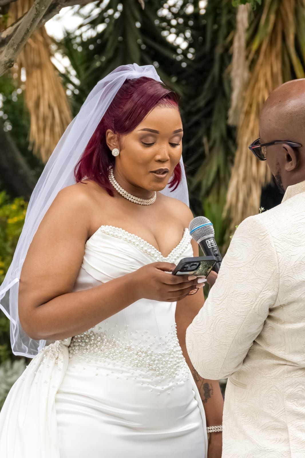Bride in white gown and veil reading vows from a phone during an outdoor wedding ceremony